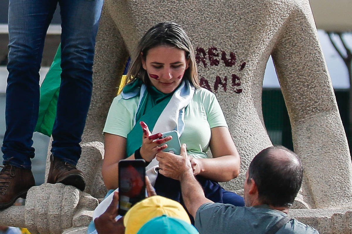 A imagem mostra uma mulher sentada em uma estátua, vestindo uma camiseta verde e segurando um celular. Ela tem marcas de tinta no rosto e está cercada por pessoas. Ao fundo, há uma bandeira do Brasil e uma inscrição na estátua que diz