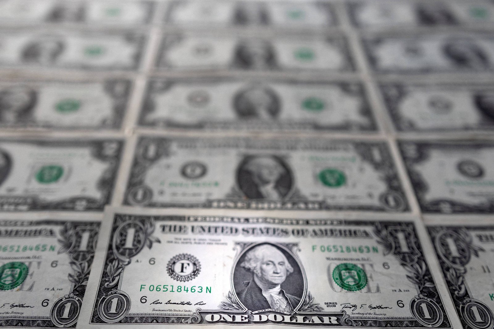 A imagem mostra várias notas de um dólar americano dispostas em uma superfície. As notas estão em uma grade, com a face de George Washington visível em cada uma delas. O fundo é desfocado, destacando as notas em primeiro plano.