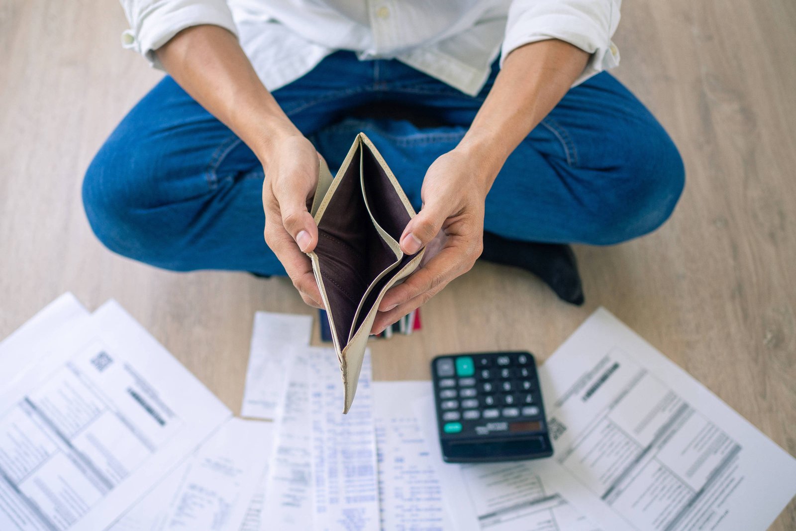 Uma pessoa sentada no chão, com as pernas cruzadas, segurando uma carteira vazia. Ao redor, há várias folhas de papel, possivelmente contas ou documentos financeiros, e uma calculadora ao lado. O ambiente parece ser uma sala com piso de madeira clara.