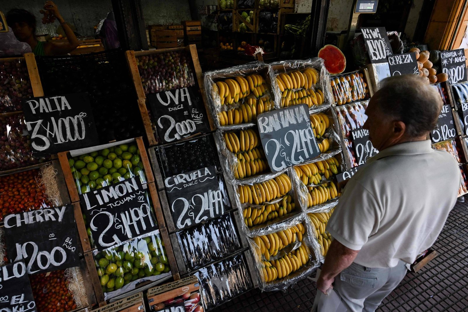 A imagem mostra um mercado de frutas com várias bancadas. À esquerda, há frutas como cerejas, limões e uvas, com preços escritos em placas pretas. À direita, há uma grande quantidade de bananas e outras frutas. Um homem de costas observa as frutas. O ambiente parece ser um mercado ao ar livre.