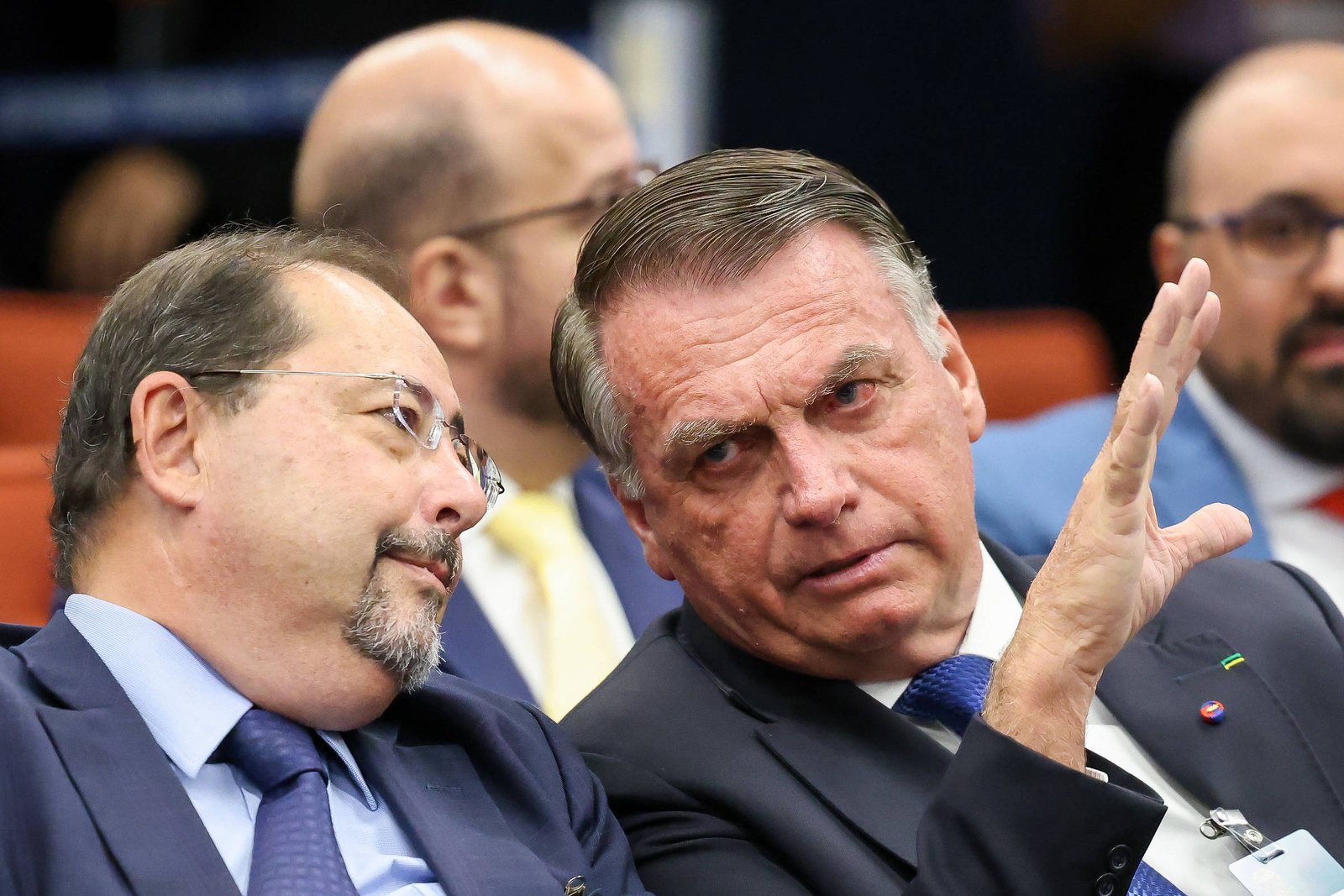 A imagem mostra dois homens conversando em um ambiente formal. Um deles está à esquerda, com óculos e um terno escuro, enquanto o outro, à direita, está com uma expressão séria e gesticulando com a mão. Ao fundo, outras pessoas estão visíveis, mas não são o foco da imagem