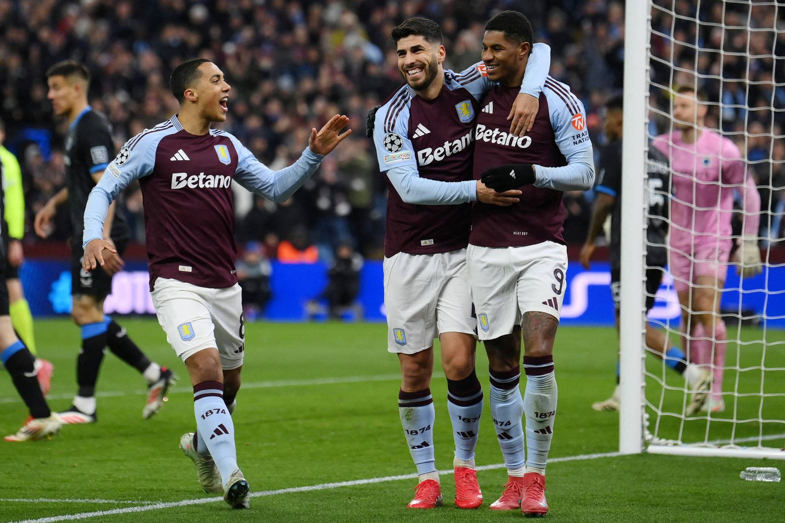 Três jogadores do Aston Villa estão celebrando em campo após um gol. Eles estão vestidos com uniformes roxos e brancos, com o logotipo da Betano visível. Ao fundo, outros jogadores estão posicionados, e um goleiro está em destaque com um uniforme rosa. O estádio está cheio de torcedores.