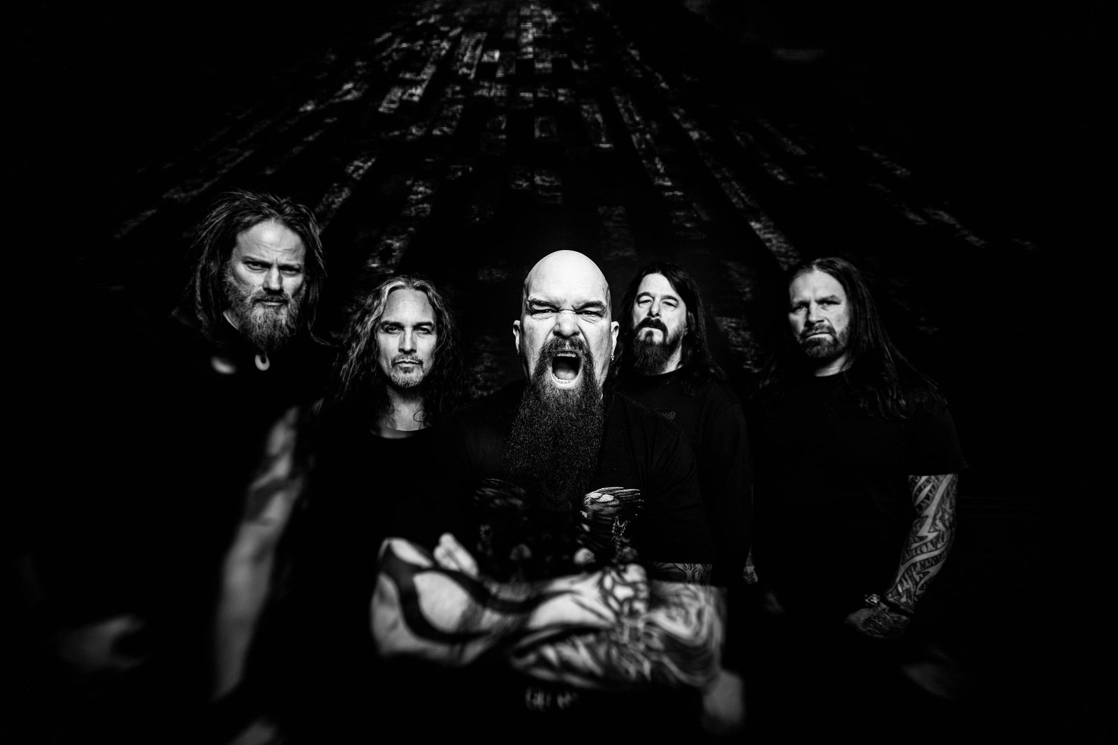 A imagem mostra cinco membros de uma banda posando juntos. Eles estão em um fundo escuro, com uma iluminação que destaca seus rostos e expressões. O vocalista, no centro, está gritando e tem uma longa barba. Os outros membros têm cabelos longos e estão vestidos com camisetas escuras. A composição é em preto e branco, criando um contraste forte.