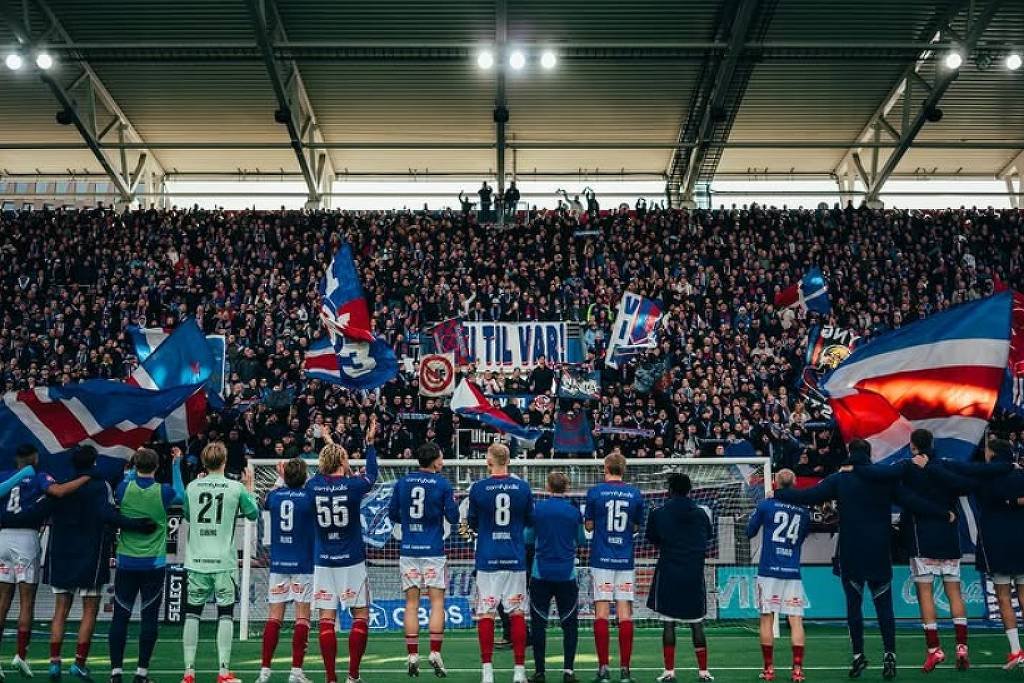 A imagem mostra uma equipe de futebol em campo, de costas, em frente a uma torcida entusiasmada. Os jogadores estão alinhados, vestindo uniformes azuis e brancos, enquanto a torcida, composta por um grande número de pessoas, segura bandeiras e faixas. O ambiente é de celebração, com luzes brilhantes e uma atmosfera vibrante.