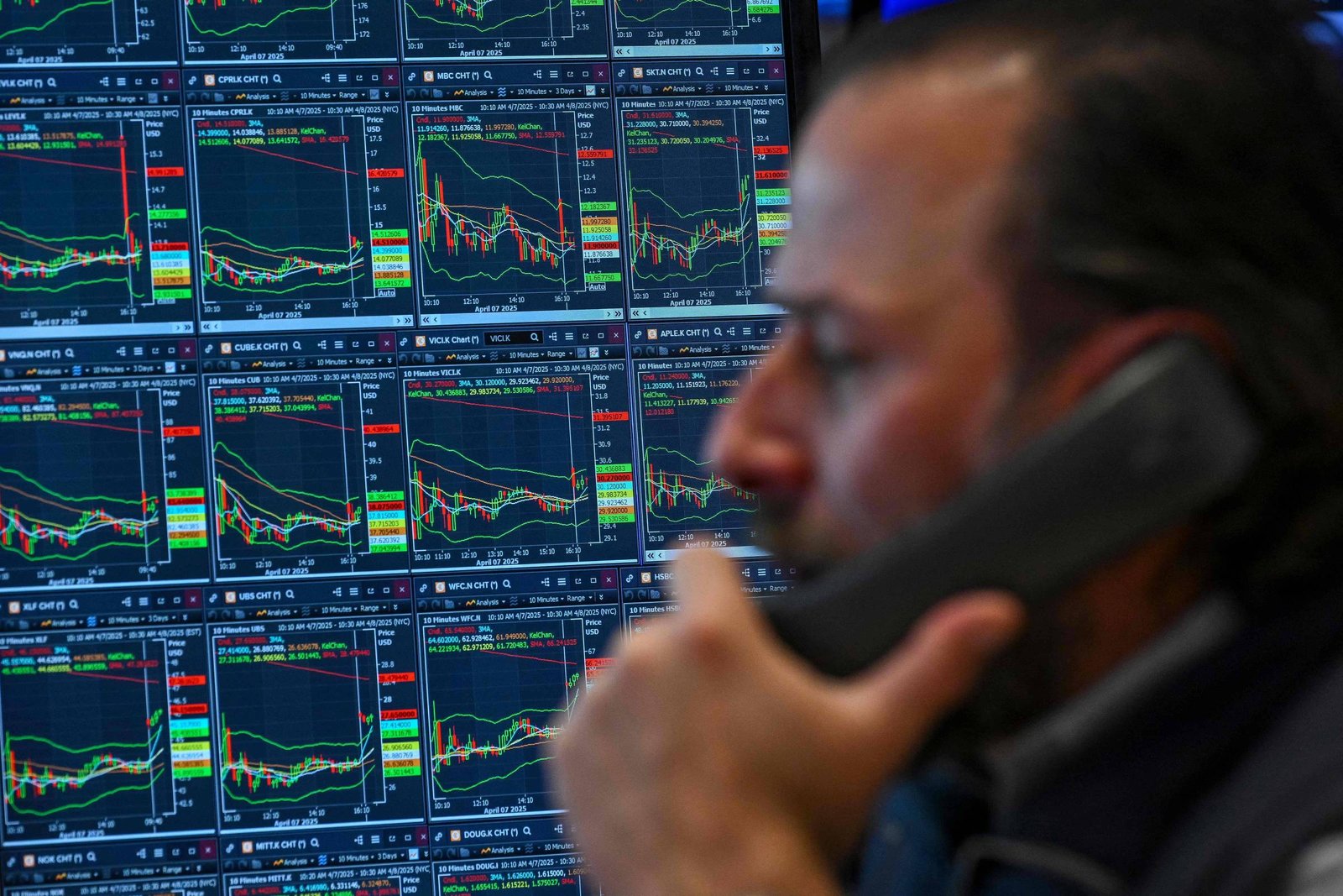 A imagem mostra um homem falando ao telefone, com um fundo de várias telas exibindo gráficos de mercado financeiro. Os gráficos apresentam linhas e barras em diferentes cores, indicando flutuações de preços. O homem está em foco, enquanto as telas atrás dele estão ligeiramente desfocadas.