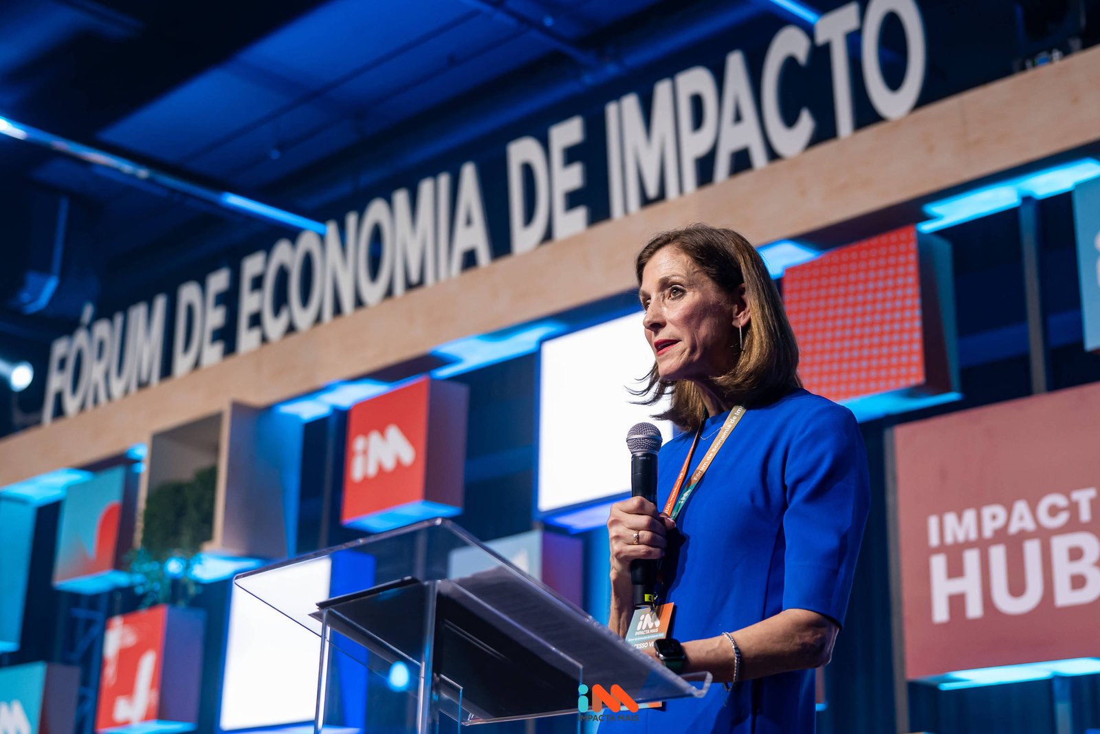 A imagem mostra uma mulher em pé atrás de um púlpito de vidro, falando em um evento. Ela usa uma blusa azul e tem cabelo solto. Ao fundo, há uma decoração com várias placas coloridas, incluindo a frase