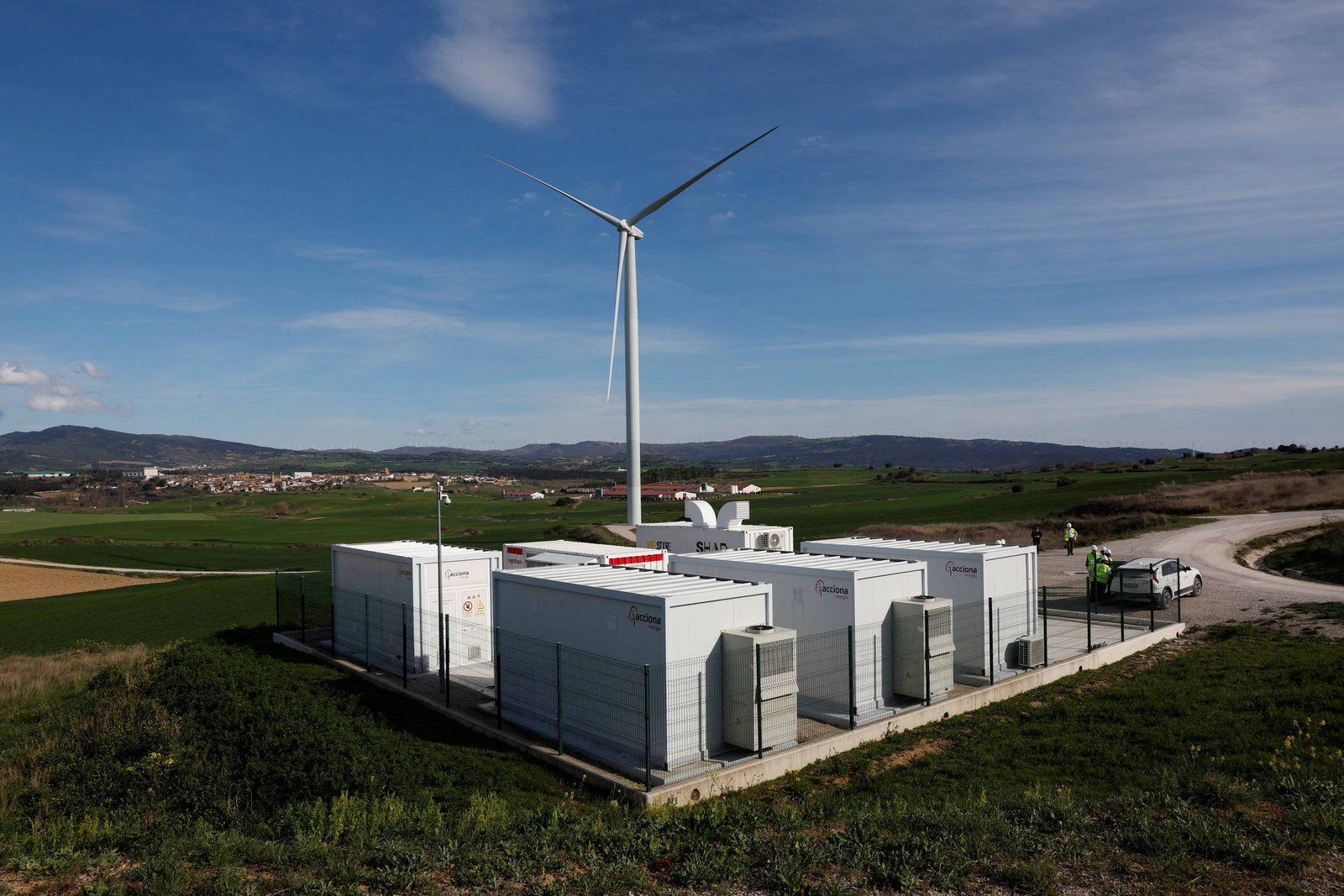 A imagem mostra uma instalação de energia renovável com um aerogerador ao fundo e várias unidades de armazenamento de energia em primeiro plano. O local está situado em uma área rural, com campos verdes ao redor e um céu azul com algumas nuvens. Há também um veículo próximo à instalação.