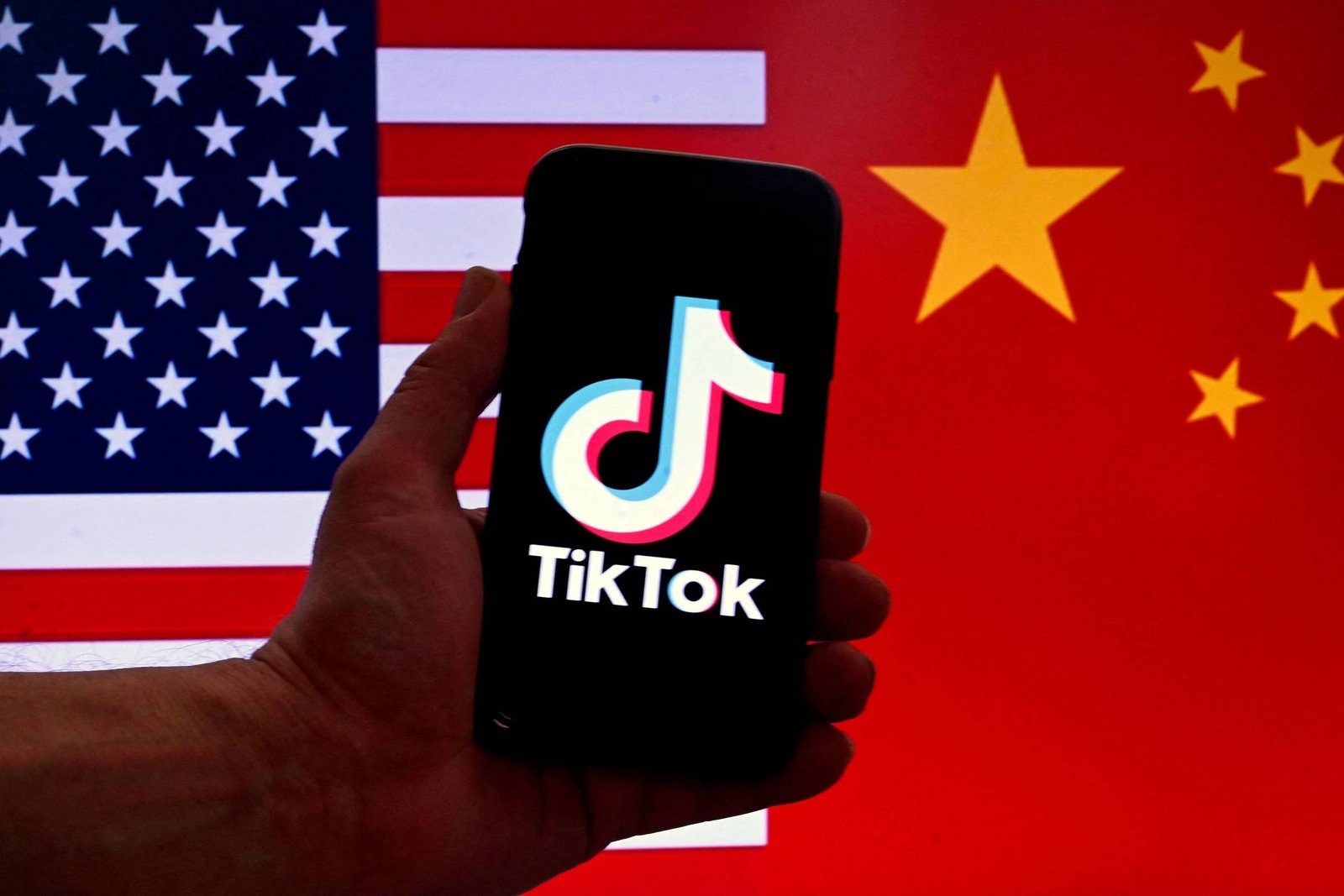A imagem mostra um smartphone segurado por uma mão, exibindo o logotipo do TikTok na tela. Ao fundo, há uma bandeira dos Estados Unidos à esquerda e uma bandeira da China à direita, criando um contraste entre os dois países.