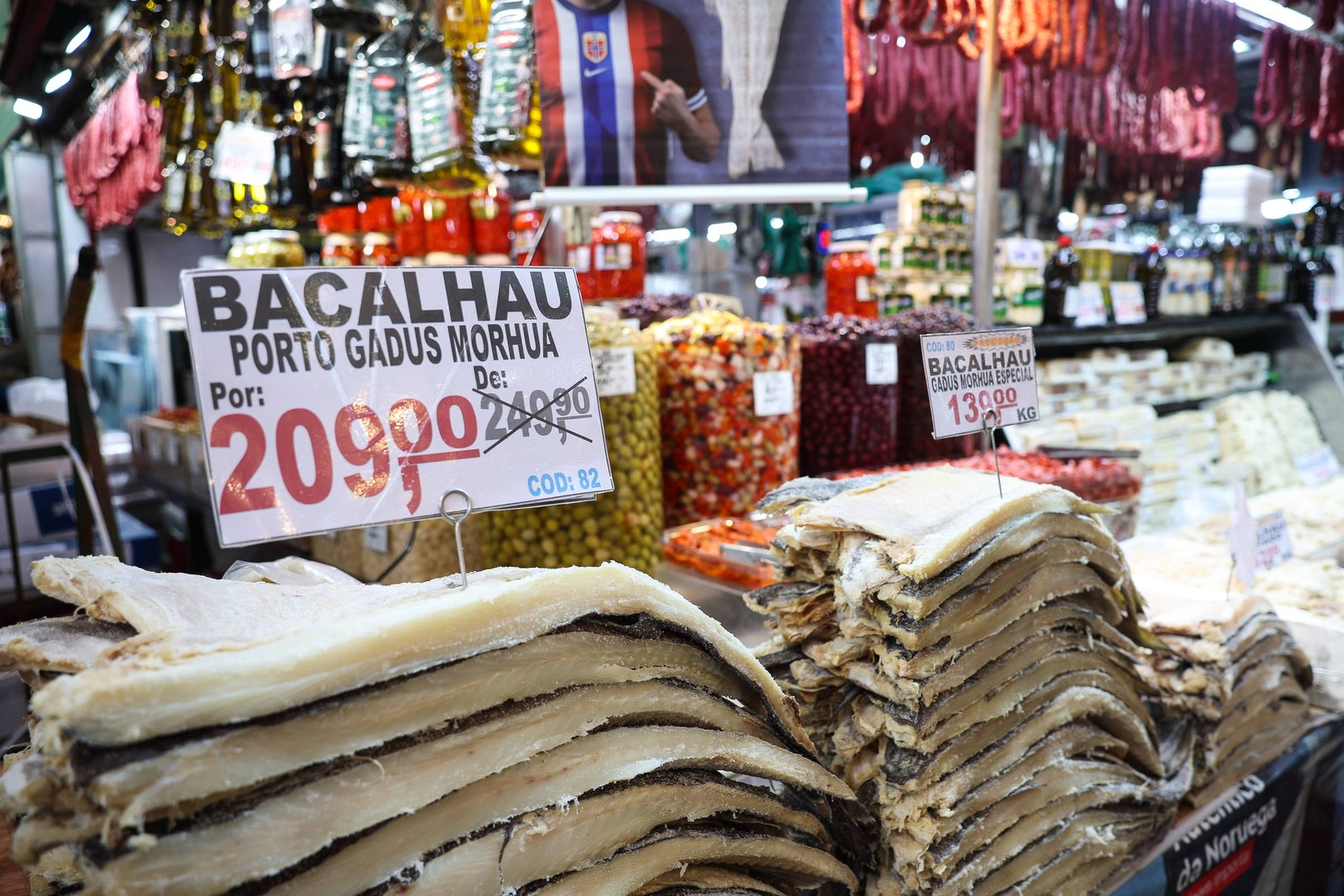 A imagem mostra uma seção de um mercado com várias mercadorias expostas. Em primeiro plano, há uma pilha de bacalhau seco, com um cartaz indicando o preço de R$ 209,90. Ao fundo, são visíveis prateleiras com outros produtos, como conservas, embutidos e temperos, além de um cartaz com o preço de R$ 139,90 para outro tipo de bacalhau. O ambiente é bem iluminado e organizado, típico de um mercado.
