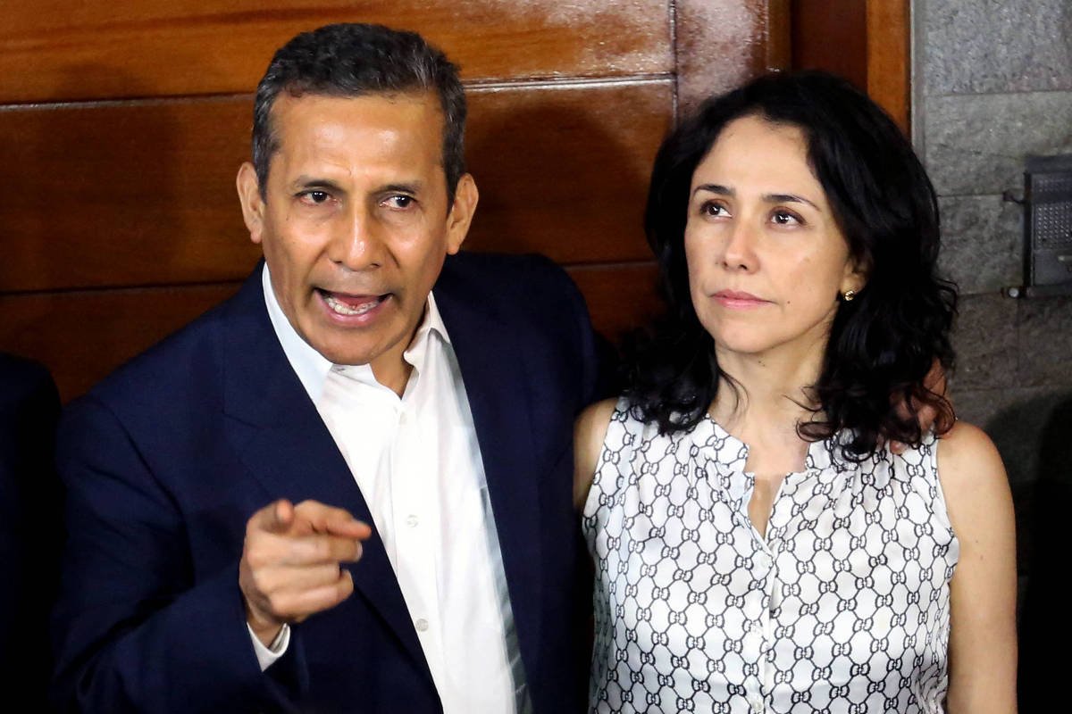 Um homem e uma mulher estão em uma conferência de imprensa. O homem, à esquerda, está gesticulando e parece estar falando de forma enfática. Ele usa um paletó escuro e uma camisa branca. A mulher, à direita, está olhando para ele e usa uma blusa branca com um padrão. O fundo é de madeira e há uma parede de pedra ao lado.