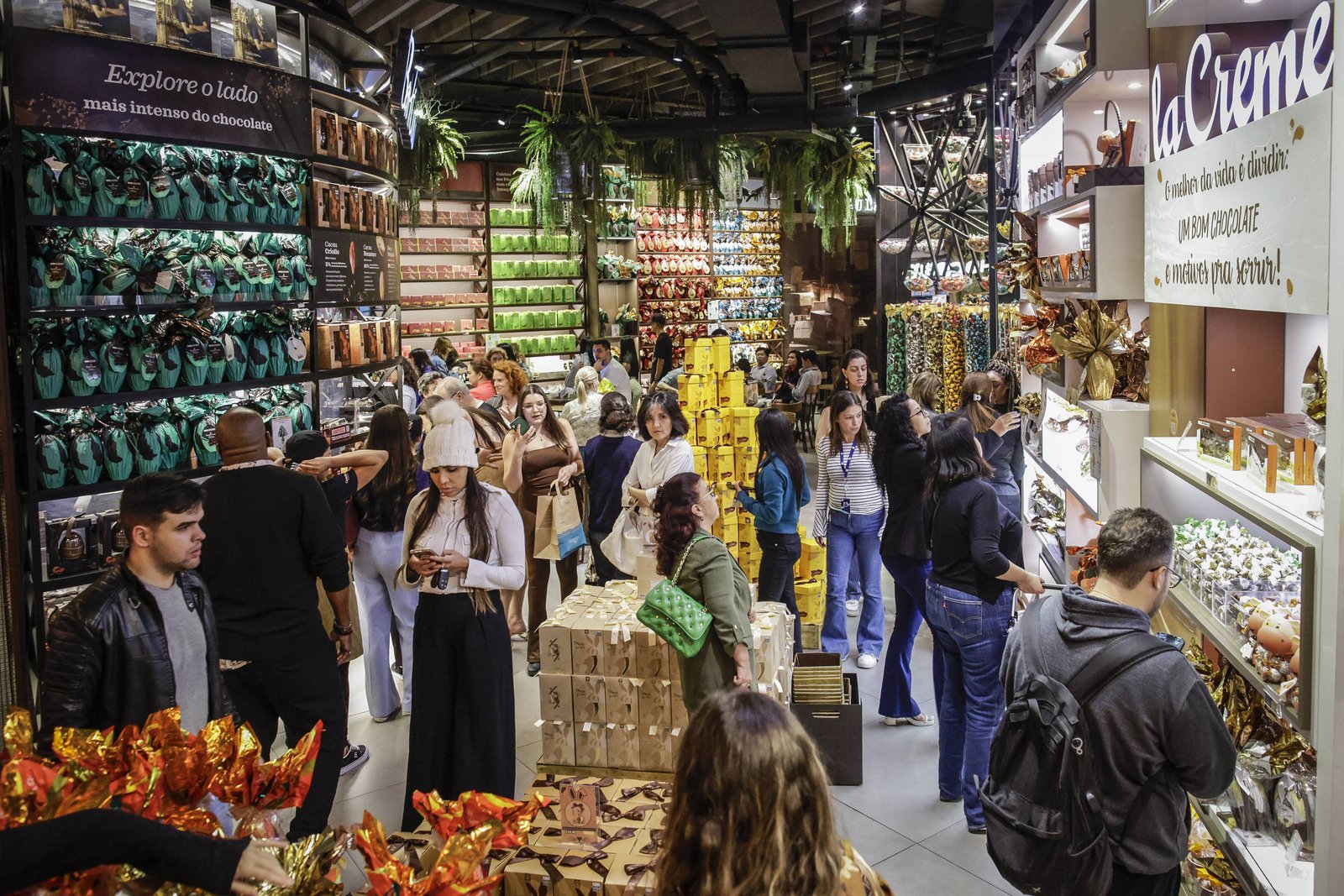 A imagem mostra um mercado interno movimentado, com várias pessoas fazendo compras. O ambiente é bem iluminado e decorado com prateleiras cheias de produtos, incluindo alimentos e itens decorativos. Há uma variedade de cores e texturas, com pessoas carregando sacolas e interagindo entre si. O espaço parece ser um local popular para compras, com uma atmosfera vibrante e cheia de vida.