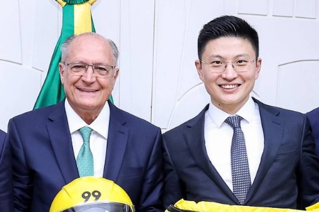 Cinco homens estão em pé em uma sala, posando para a foto. No centro, um homem mais velho, aparentemente um político, segura um capacete amarelo com o número 99. À sua esquerda, um homem de terno escuro e gravata clara segura uma bolsa amarela com o número 99. Os outros três homens estão ao redor, todos vestidos formalmente. Ao fundo, há uma bandeira do Brasil e uma parede branca.