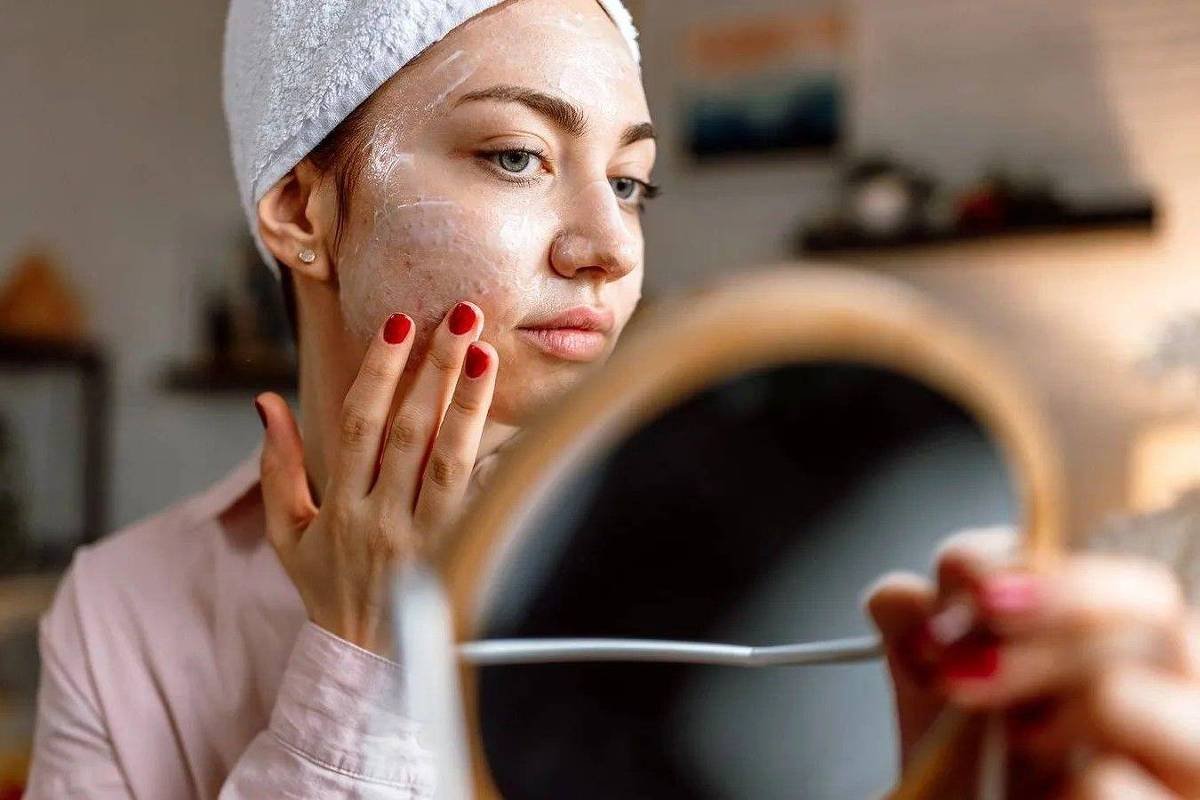 Uma mulher está olhando para um espelho enquanto aplica uma máscara facial em seu rosto. Ela usa uma toalha na cabeça e está vestida com uma camisa de botão. O ambiente ao fundo é bem iluminado, com prateleiras e objetos decorativos visíveis.