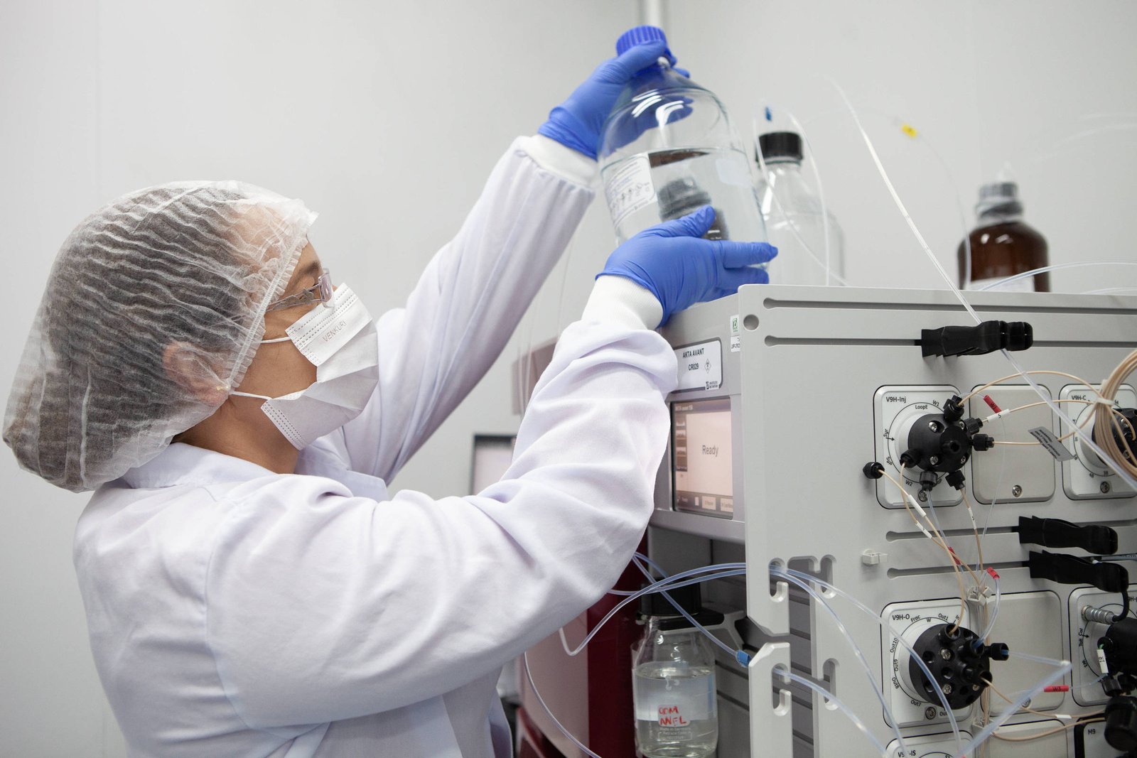 Uma profissional de laboratório, usando um jaleco branco, luvas azuis e máscara, está segurando um frasco grande enquanto o coloca em um equipamento de laboratório. O ambiente é limpo e organizado, com vários frascos e tubos visíveis ao fundo.
