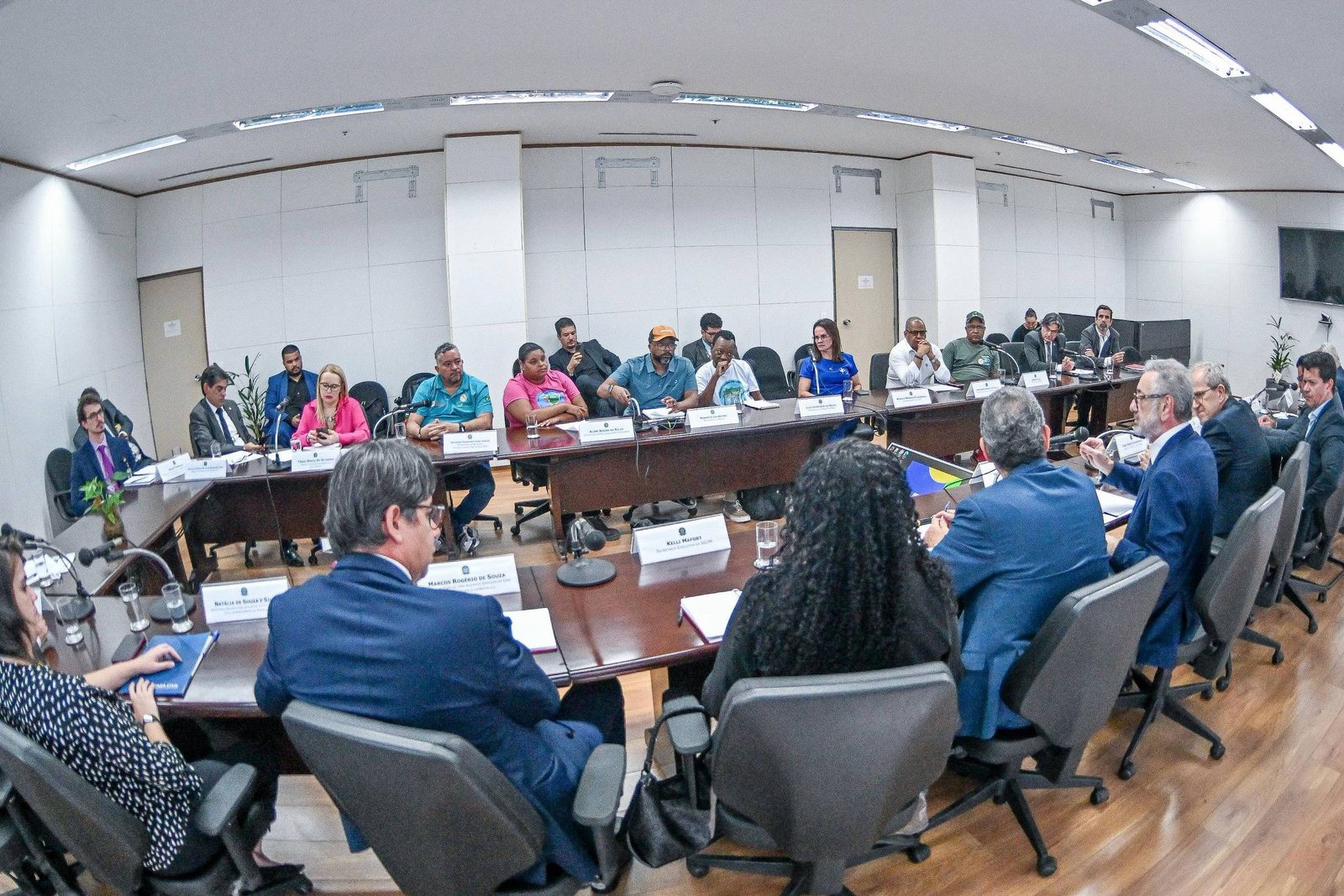 A imagem mostra uma sala de reuniões com várias pessoas sentadas ao redor de uma mesa. Há um grupo de pessoas em primeiro plano, algumas delas de costas, e outras em segundo plano, participando da discussão. A sala possui paredes brancas e um piso de madeira, com janelas que permitem a entrada de luz natural. Em uma das paredes, há uma tela de televisão.