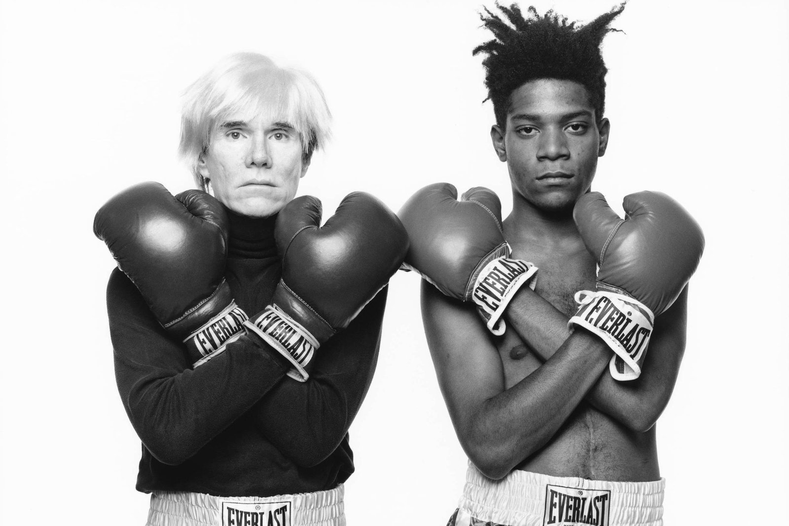 A imagem em preto e branco mostra duas figuras masculinas vestindo calças de boxe prateadas e luvas de boxe. A figura à esquerda tem cabelo platinado e usa uma blusa preta, enquanto a figura à direita tem cabelo crespo e está sem camisa. Ambos estão em uma pose de luta, com os braços cruzados e as luvas levantadas.