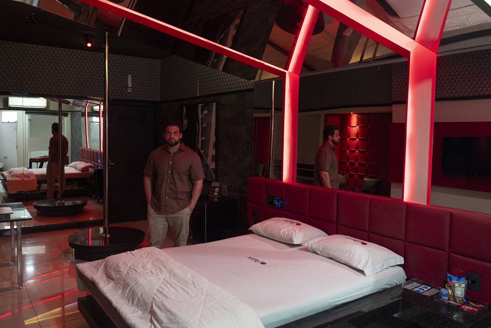 A imagem mostra um quarto moderno com iluminação vermelha. No centro, há uma cama de casal com lençóis brancos. Um homem está em pé ao lado da cama, e há um espelho refletindo o ambiente. O quarto possui paredes com texturas e um design contemporâneo, incluindo uma mesa de vidro e uma televisão na parede.