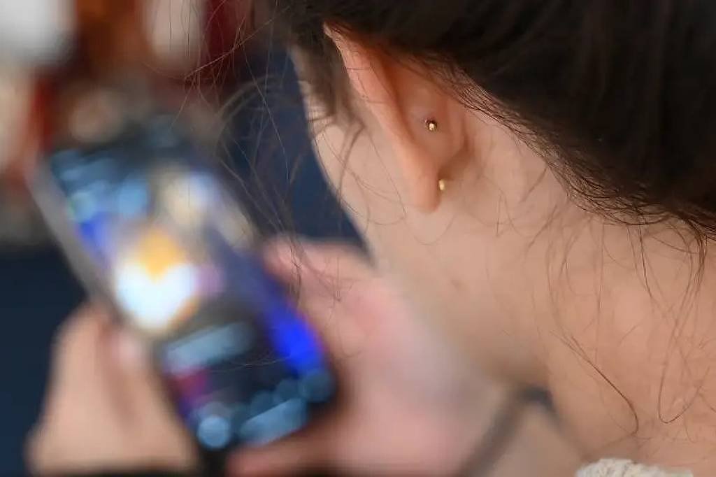 A imagem mostra a parte de trás da cabeça de uma pessoa com cabelo castanho escuro, que está segurando um smartphone. O foco está no telefone, que exibe uma tela iluminada, enquanto o fundo é desfocado. A pessoa parece estar concentrada no que está vendo no dispositivo.