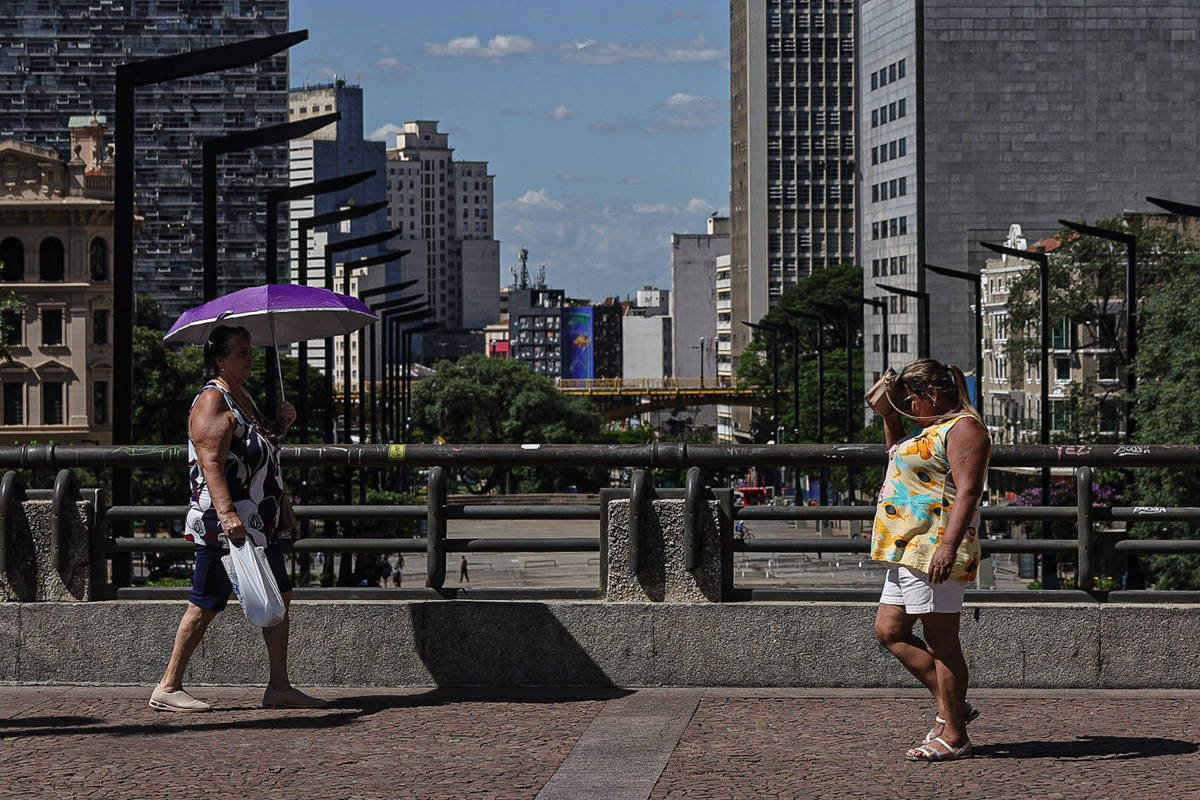 Duas mulheres caminham em uma calçada em uma área urbana. Uma mulher usa um guarda-chuva roxo e carrega uma sacola, enquanto a outra mulher, vestindo uma blusa amarela e shorts brancos, parece estar se protegendo do sol. Ao fundo, há prédios altos e um céu azul com algumas nuvens.