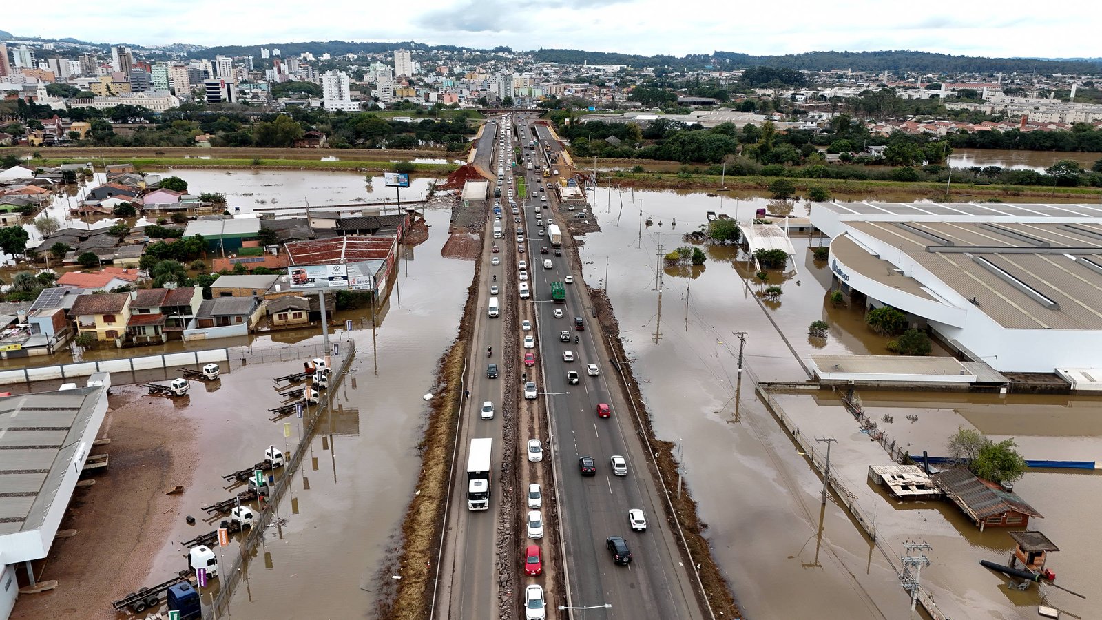 A imagem mostra uma estrada em uma área urbana, com veículos se movendo em uma pista central. À esquerda e à direita da estrada, há água acumulada devido a uma inundação, cobrindo parte das casas e estabelecimentos próximos. Ao fundo, é possível ver prédios e uma área urbana mais extensa, com vegetação ao redor. O céu está nublado, indicando um clima chuvoso.