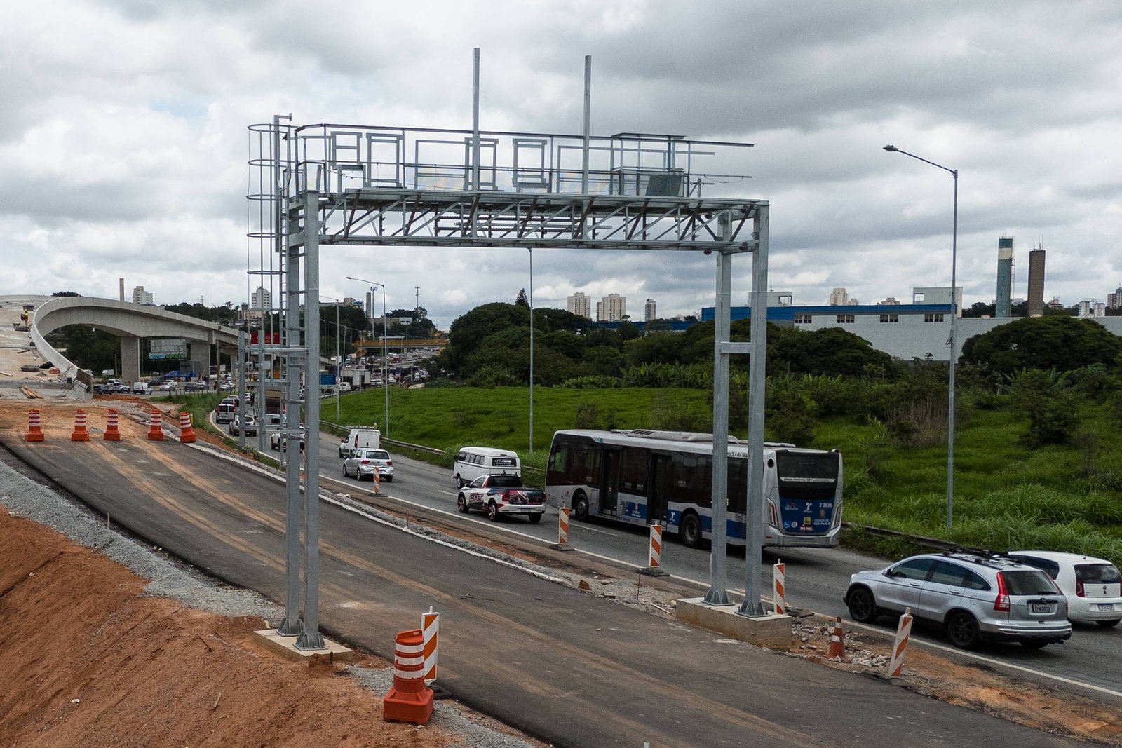 A imagem mostra uma estrada com sinalização e obras ao lado. Há uma estrutura de pedágio free flow e vários veículos, incluindo um ônibus e carros, parados na via. O céu está nublado e há áreas de grama ao fundo, além de estruturas industriais visíveis.