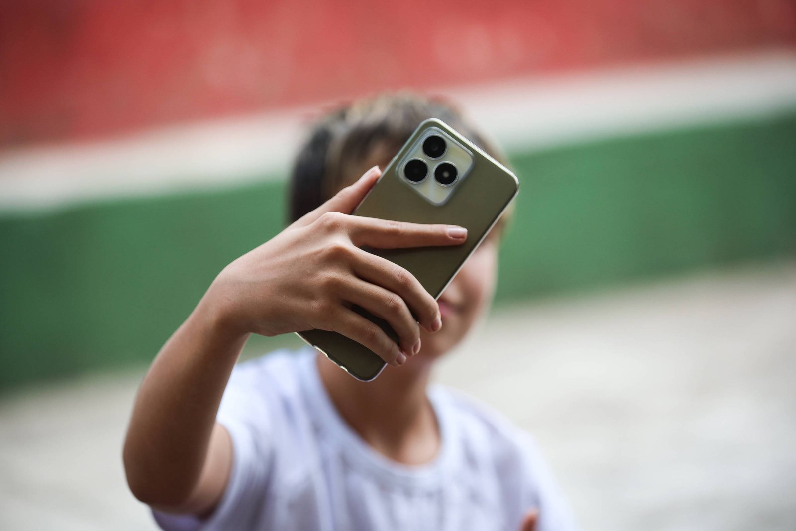 Uma criança está tirando uma selfie com um smartphone. A criança segura o telefone com uma mão, enquanto a outra mão está visível. O fundo apresenta uma parede com cores vermelha e verde, sugerindo um ambiente externo ou um espaço público.