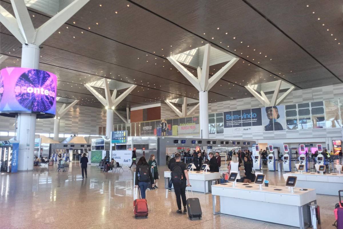 A imagem mostra o interior de um aeroporto moderno, com um grande espaço aberto e tetos altos. Há várias pessoas se movendo com malas, algumas paradas em balcões de check-in. O piso é de cerâmica clara e há colunas estruturais visíveis. Ao fundo, há um grande painel digital com cores vibrantes.