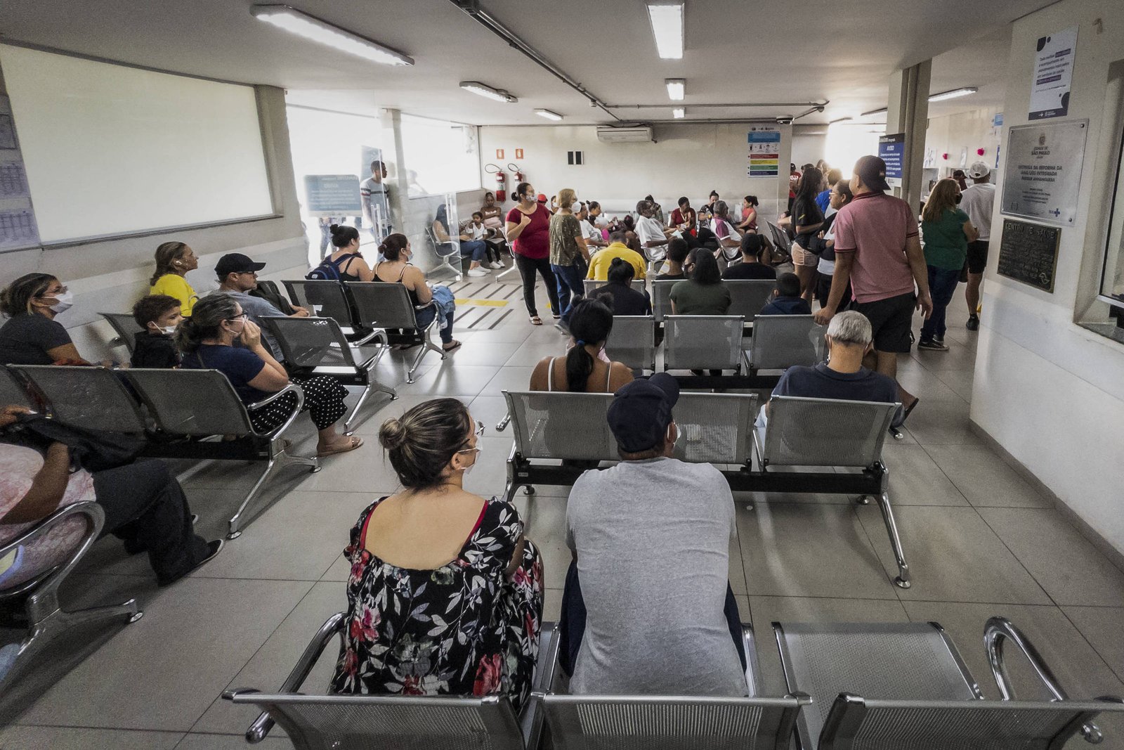 A imagem mostra uma sala de espera em um hospital, com várias pessoas sentadas em cadeiras de metal. Algumas pessoas estão usando máscaras. O ambiente é bem iluminado, com paredes brancas e piso de cerâmica. Ao fundo, há um balcão de atendimento e um grupo de pessoas em pé, possivelmente aguardando atendimento. A sala parece estar cheia, com um número considerável de pacientes esperando.