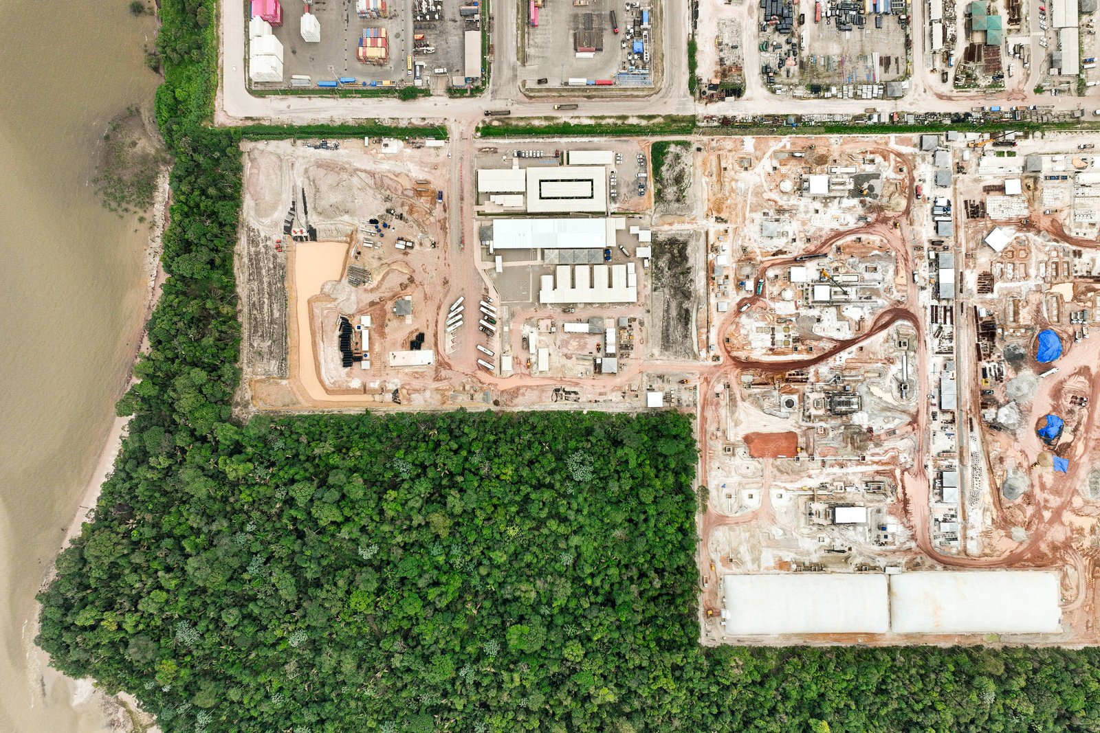 A imagem mostra uma vista aérea de uma área de construção adjacente a uma floresta densa. À esquerda, há uma faixa de vegetação verde, enquanto à direita, várias estruturas em construção são visíveis, incluindo edifícios e áreas de terraplanagem. O solo é predominantemente marrom, indicando atividade de construção. No canto inferior esquerdo, pode-se ver um corpo d