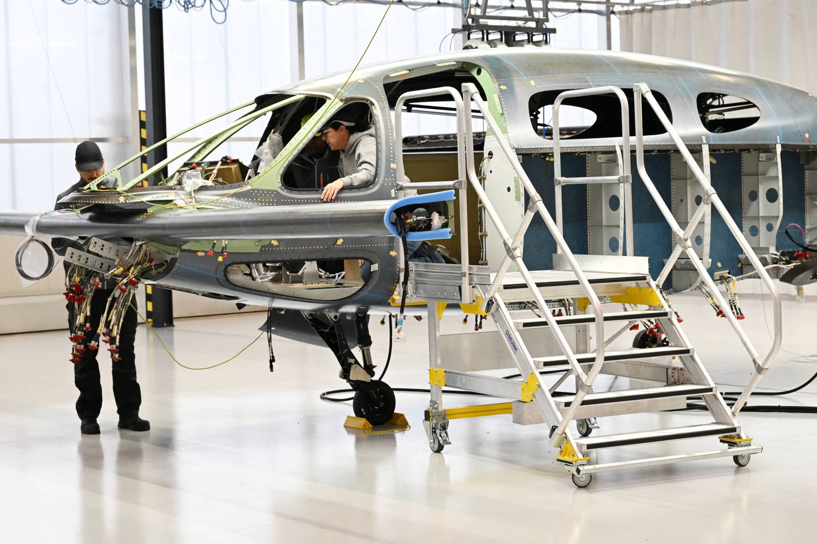 A imagem mostra um ambiente de oficina onde uma aeronave está em processo de montagem. Um trabalhador está posicionado dentro da fuselagem da aeronave, enquanto outro trabalhador está ao lado, observando. A estrutura da aeronave é visível, com partes metálicas expostas e escadas de acesso ao lado. O ambiente é bem iluminado, com janelas grandes ao fundo.
