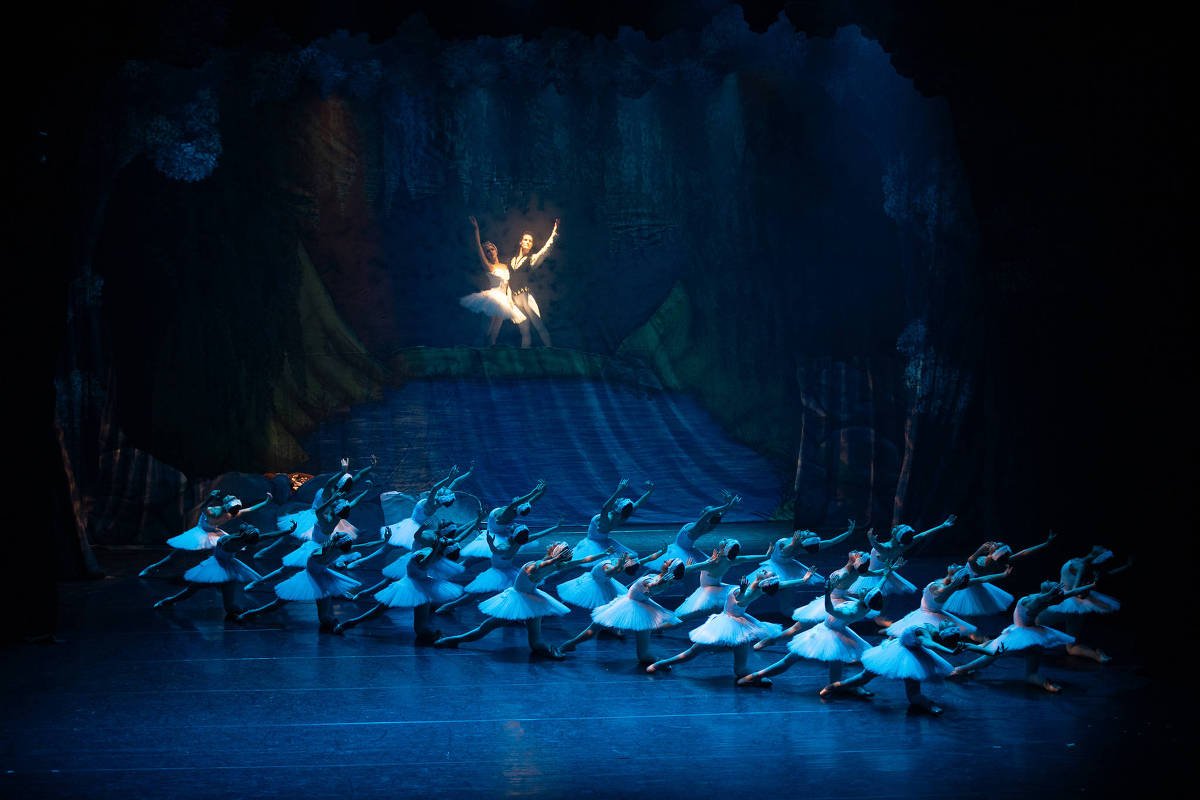 A imagem mostra um grupo de bailarinas em um palco, todas vestindo saias brancas e dançando em sincronia. No fundo, uma bailarina se destaca, flutuando no ar, iluminada por um foco de luz. O cenário é escuro, com elementos que sugerem um ambiente natural, como árvores e água.