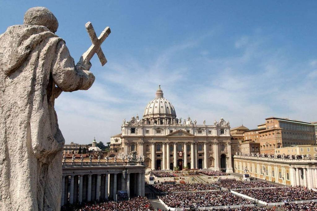 A imagem mostra a Praça de São Pedro no Vaticano, com uma grande multidão reunida. Em primeiro plano, há uma estátua de um santo segurando uma cruz. Ao fundo, destaca-se a Basílica de São Pedro, com sua cúpula imponente e arquitetura clássica. O céu está claro e ensolarado, contribuindo para a atmosfera vibrante do local.