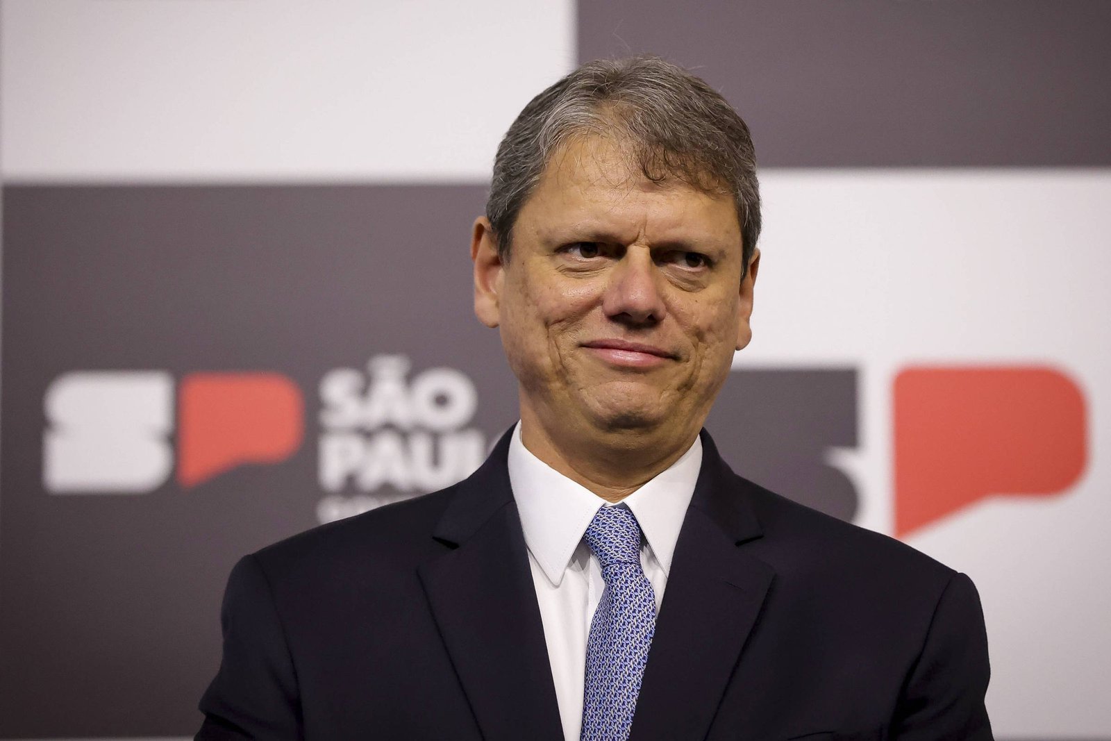 Um homem com cabelo grisalho e liso, usando um terno escuro e uma gravata azul, está sorrindo levemente. Ao fundo, há um painel com as palavras
