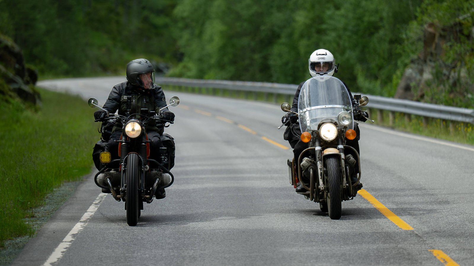 A imagem mostra dois motociclistas em uma estrada. O motociclista à esquerda está em uma moto preta e usa um capacete escuro, enquanto o motociclista à direita está em uma moto de estilo clássico e usa um capacete branco. A estrada é cercada por vegetação verde ao lado.