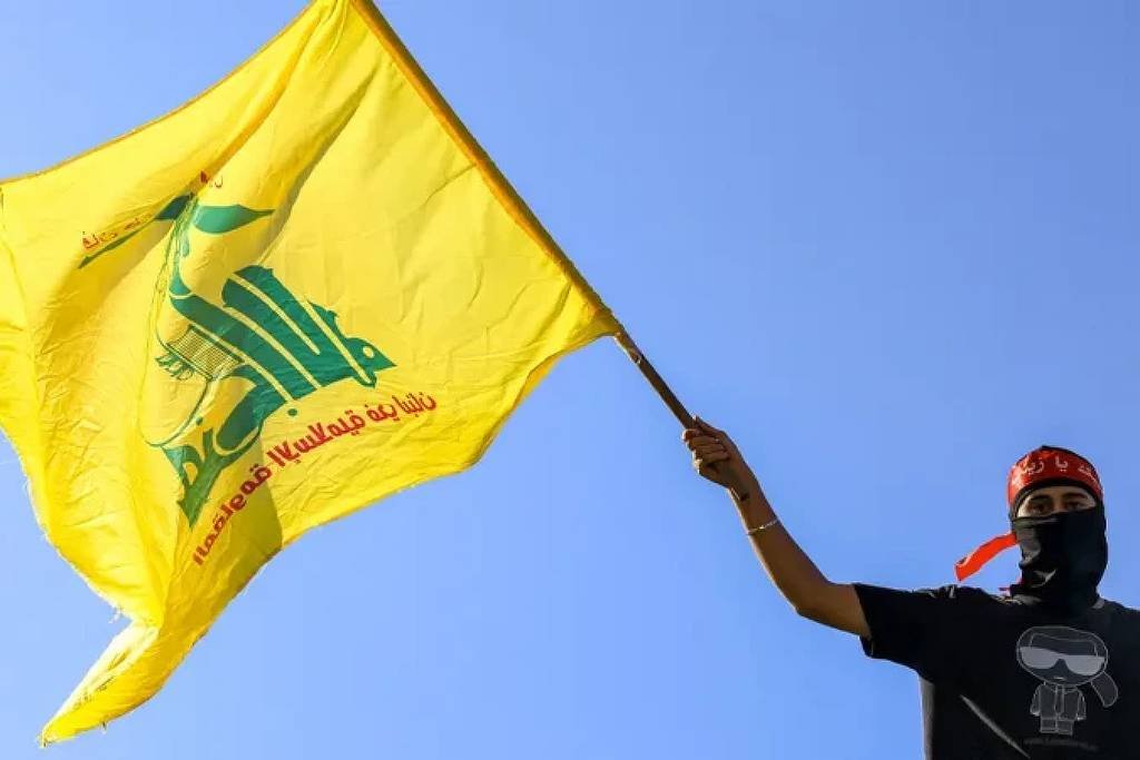 Um homem está segurando uma bandeira amarela com inscrições em árabe. Ele está usando uma máscara preta e uma camiseta escura. O fundo é um céu azul claro.