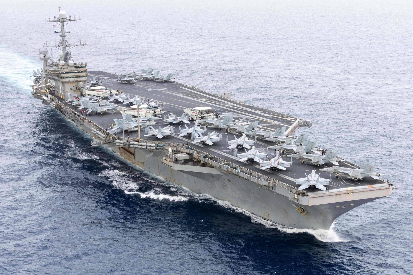 A imagem mostra um porta-aviões navegando em águas abertas. O navio possui uma pista de decolagem visível na parte superior, onde estão estacionados vários aviões de combate. O céu está nublado e o mar parece calmo, com pequenas ondas.