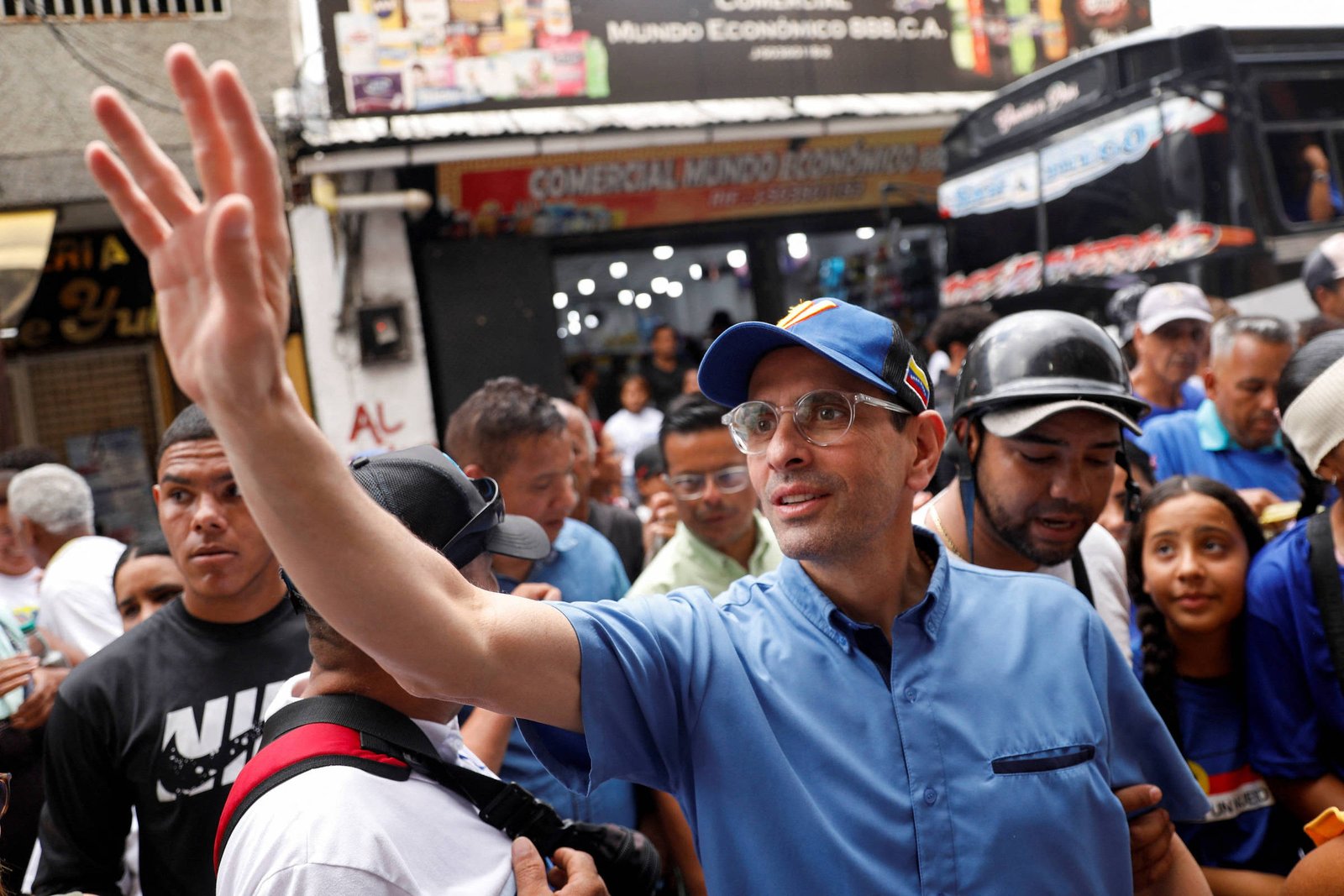 A imagem mostra um grupo de pessoas em uma rua movimentada. No centro, um homem com óculos e um boné azul acena com a mão. Ele está vestido com uma camisa azul e parece estar interagindo com a multidão ao seu redor. Ao fundo, há várias pessoas, algumas usando camisetas de marcas conhecidas, e uma loja com letreiros visíveis. O ambiente é urbano e parece haver um clima de agitação.