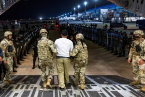 A imagem mostra um grupo de soldados em uniformes camuflados, acompanhando um homem que está sendo levado para dentro de uma aeronave militar. O ambiente é noturno e há uma formação de policiais em pé ao fundo, alinhados em uma fila. A iluminação do aeroporto é visível, com luzes brilhantes ao fundo, e a cena parece ser uma operação de segurança ou transporte de prisioneiro.