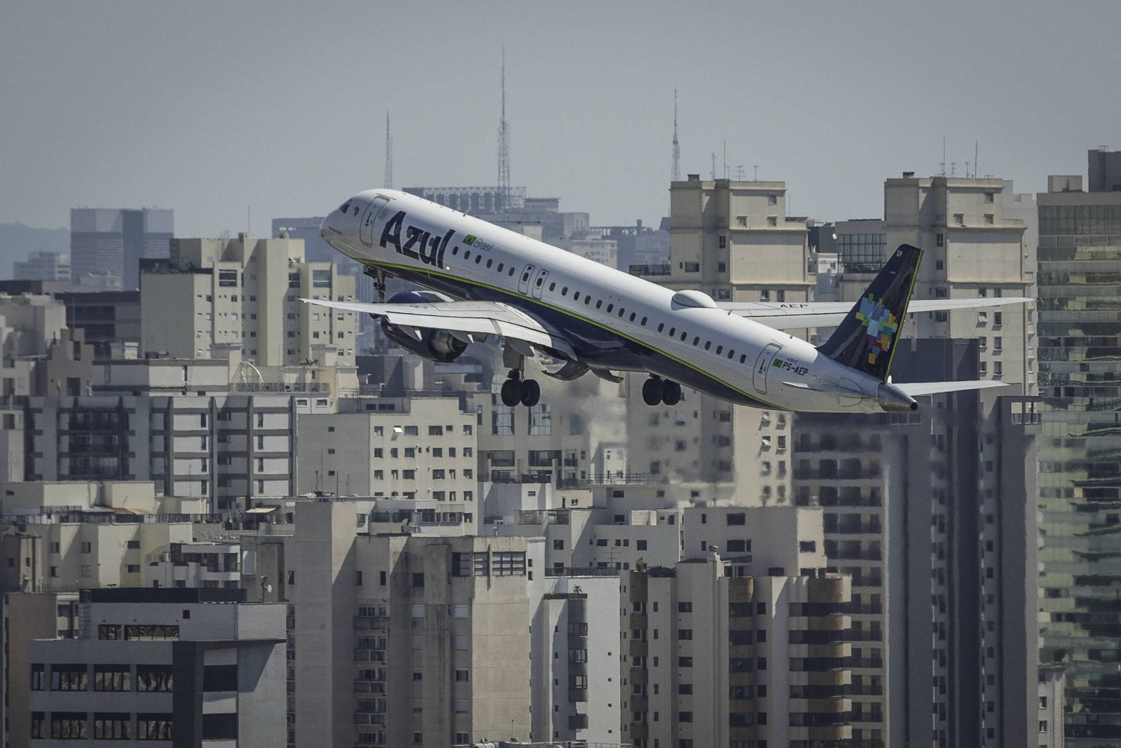 Um avião está decolando em direção ao céu, com a cidade ao fundo. Os edifícios altos e modernos são visíveis, formando um cenário urbano denso. O céu está claro, e o avião é de uma companhia aérea brasileira, com a fuselagem predominantemente branca e detalhes em azul e verde.