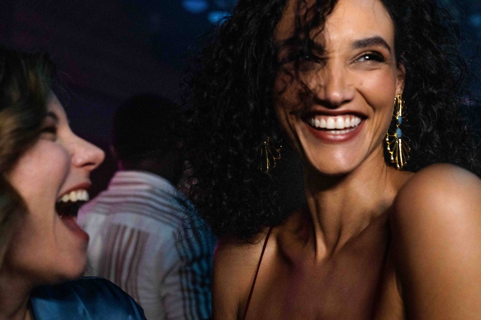 A imagem mostra duas mulheres sorrindo em um ambiente de festa. A mulher à esquerda tem cabelo castanho e ondulado, vestindo uma blusa azul. A mulher à direita tem cabelo cacheado e usa brincos grandes, sorrindo amplamente. Ao fundo, há pessoas desfocadas e luzes coloridas, sugerindo um ambiente festivo.