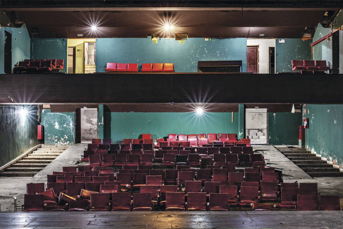 A imagem mostra o interior de um teatro abandonado, com assentos vermelhos dispostos em várias fileiras. O palco está visível na parte inferior, e há uma área superior com mais assentos. As paredes são de um tom verde desgastado, e a iluminação é fraca, com algumas luzes no teto. O ambiente parece estar em estado de deterioração, com marcas e sujeira visíveis nas paredes e no chão.