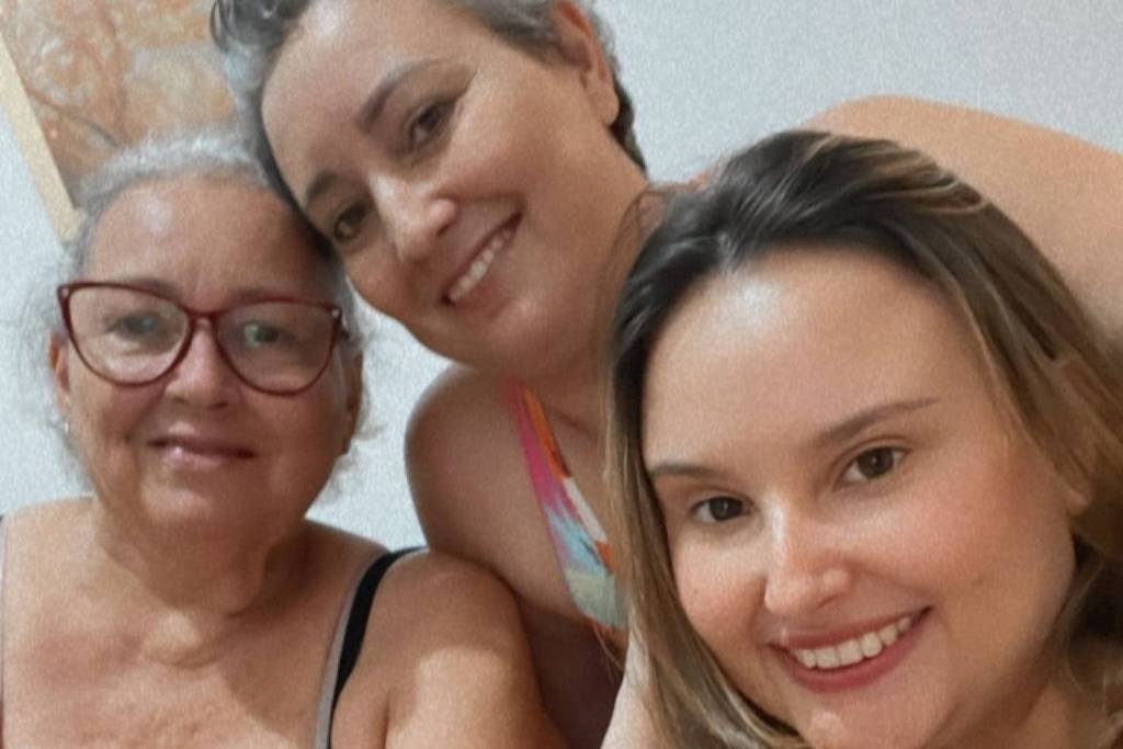 A imagem mostra três mulheres sorrindo para a câmera. A mulher à esquerda é mais velha, com cabelo grisalho e óculos, vestindo uma blusa listrada. A mulher do meio tem cabelo curto e liso, e está usando uma blusa colorida. A mulher à direita é mais jovem, com cabelo longo e liso, e está usando uma blusa preta. Elas estão em um ambiente interno, com uma parede clara ao fundo.