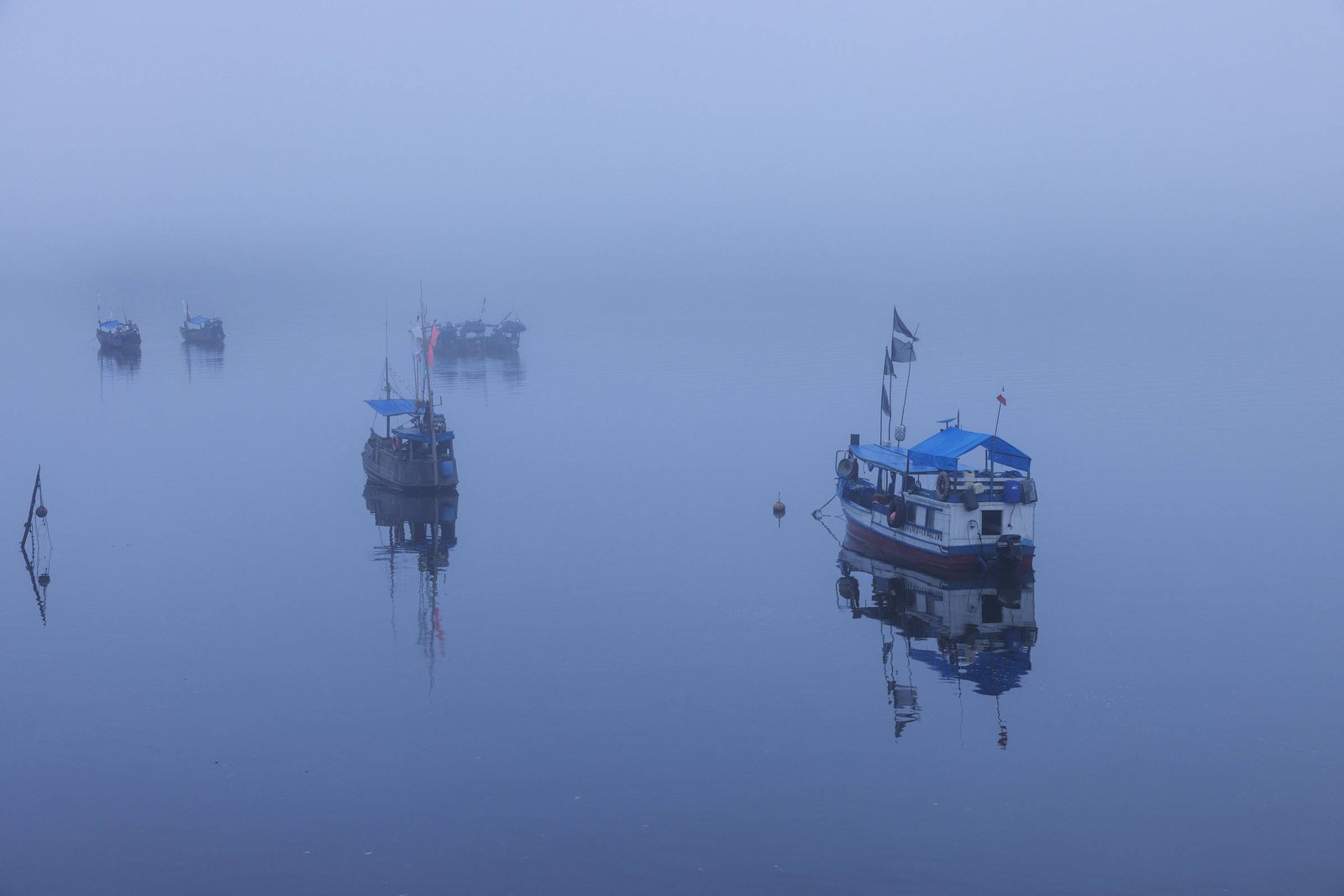A imagem mostra vários barcos ancorados. Os barcos, de diferentes tamanhos, estão refletidos na água calma, criando um efeito de espelho. O ambiente é predominantemente cinza
