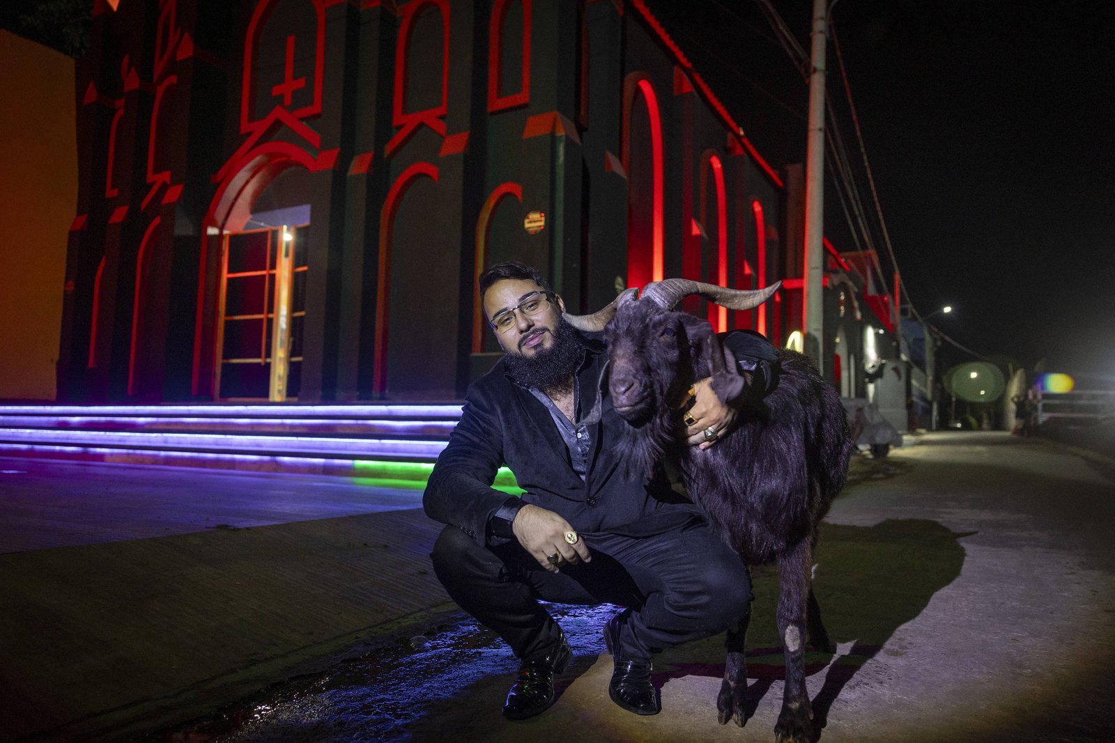 Um homem está agachado ao lado de uma cabra em uma rua iluminada à noite. O homem usa um traje escuro e óculos, enquanto a cabra está próxima a ele. Ao fundo, há um edifício com iluminação colorida, destacando-se em vermelho e azul. A cena é noturna, com iluminação artificial visível.