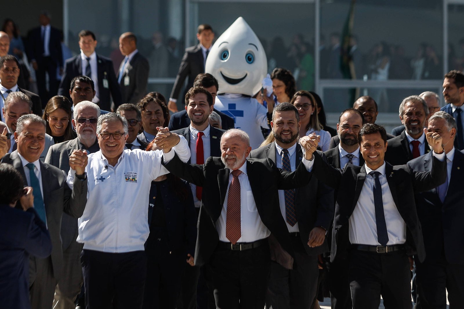 Um grupo de pessoas posando para uma foto em um evento ao ar livre. No centro, um homem de terno escuro e gravata vermelha segura as mãos de outros participantes. Ao fundo, um mascote com uma aparência amigável, representando uma gota d