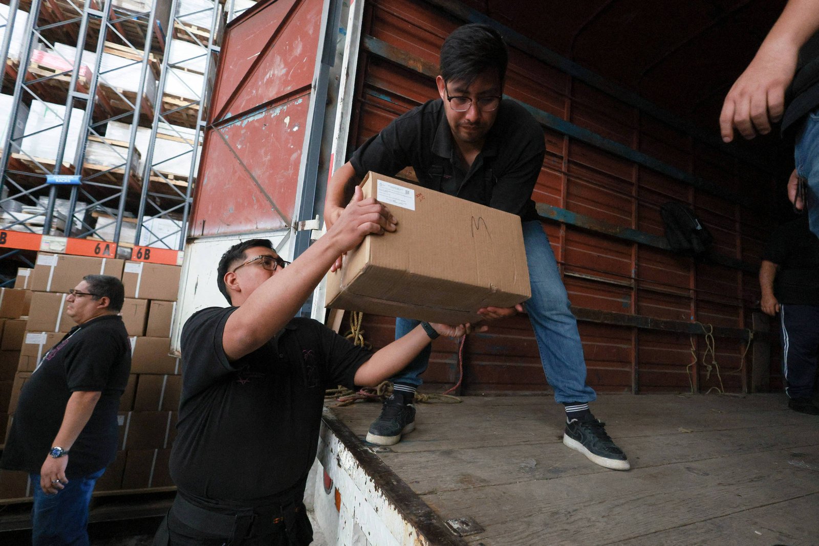 Um grupo de pessoas, possivelmente trabalhadores, envolvidos no processo de descarregamento de caixas. Estão vestidos com roupas de trabalho, predominantemente pretas e cinzas. O ambiente é um armazém. A estrutura de armazém de estantes, com outros caminhões ou estruturas e mercadorias