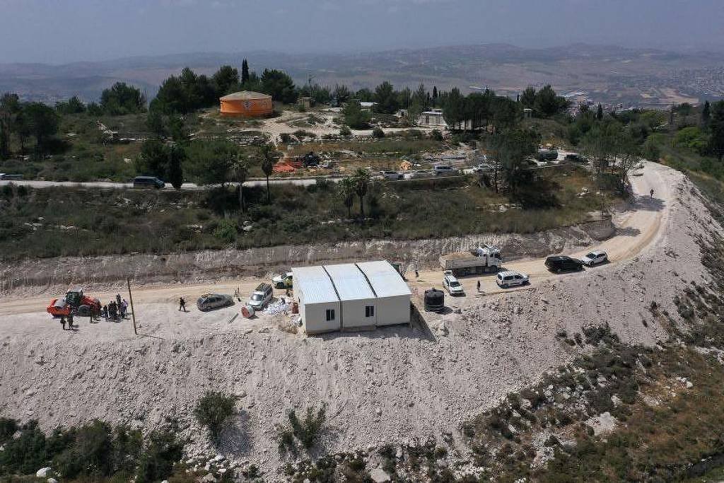 A imagem mostra uma vista aérea de uma área rural com uma estrada de terra. No primeiro plano, há uma pequena construção branca, possivelmente um escritório ou abrigo, cercada por veículos estacionados. Ao fundo, há uma estrutura laranja e várias árvores. A paisagem é montanhosa, com uma vista ampla da região ao redor.