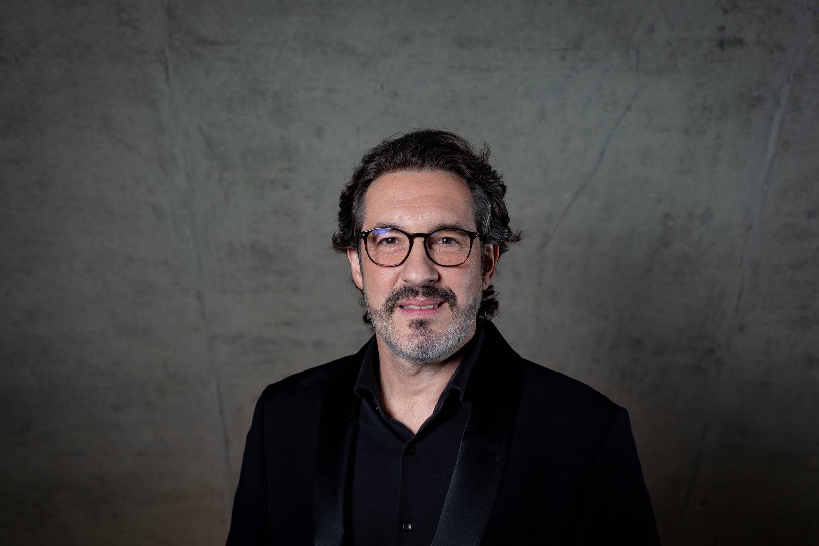 Um homem com cabelo escuro e ondulado, usando óculos e um terno preto, está posando para a foto. O fundo é uma parede de concreto com uma textura suave e neutra. O homem tem uma expressão amigável e está olhando diretamente para a câmera.