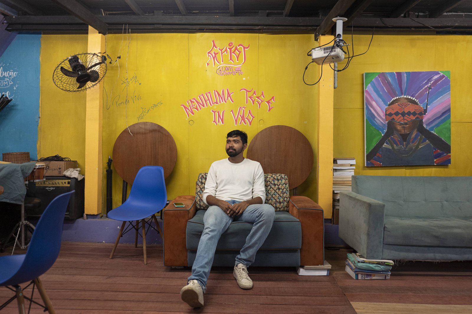A imagem mostra um homem sentado em um sofá em um ambiente colorido. As paredes são amarelas e há uma arte em uma das paredes, além de um quadro com uma figura feminina. O homem está vestido com uma camiseta clara e jeans, e parece relaxado. No fundo, há ventiladores de teto e algumas cadeiras azuis. O chão é de madeira e há livros empilhados ao lado do sofá.