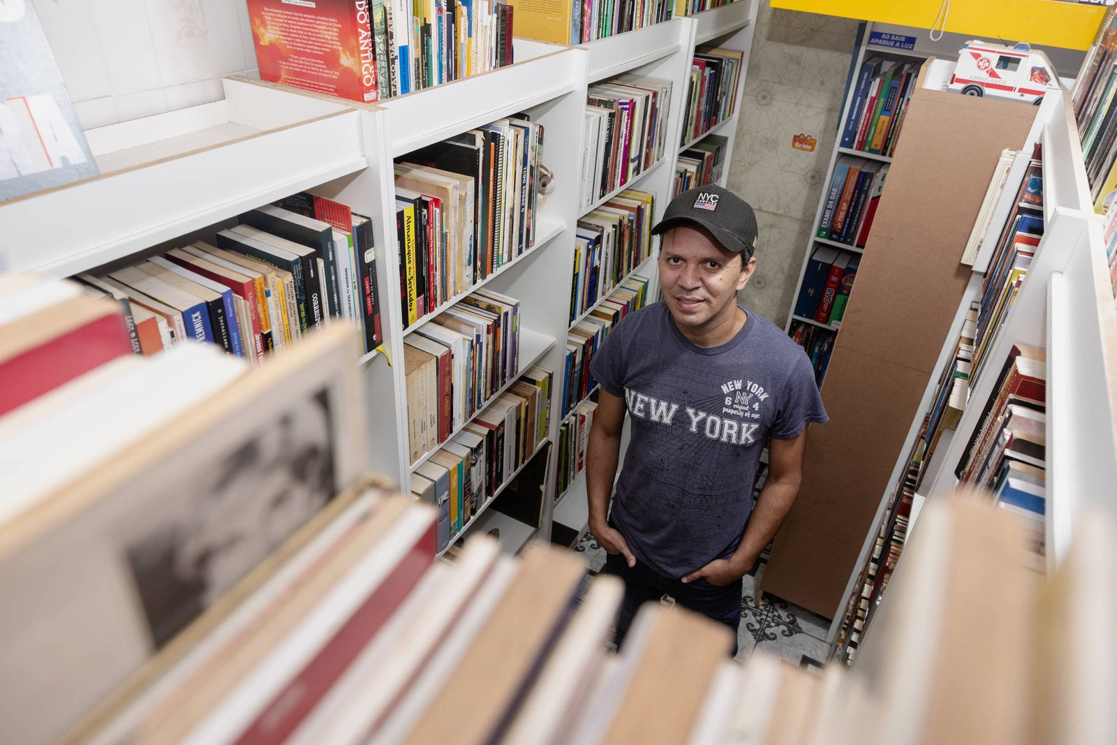 Um homem está em uma biblioteca, cercado por estantes cheias de livros. Ele usa uma camiseta cinza com a palavra